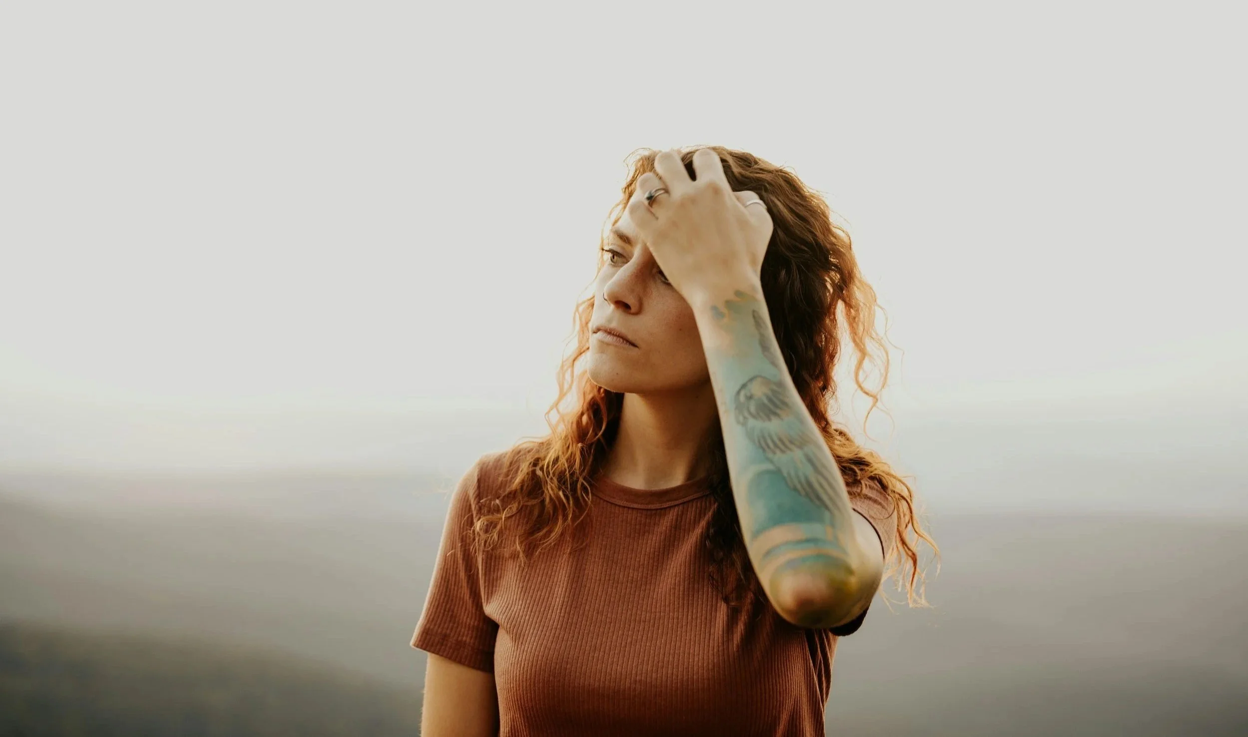 A woman standing outside looking into the distance running a hand through her hair wearing a brown t-shirt with forearm tattoos.