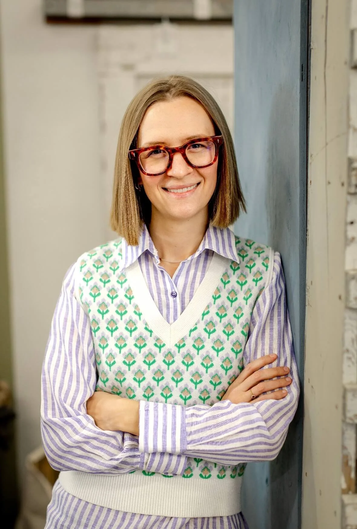 Woman with shoulder-length brown hair, wearing red tortoiseshell glasses, a striped shirt, and a white vest with green floral patterns, standing with arms crossed and smiling.