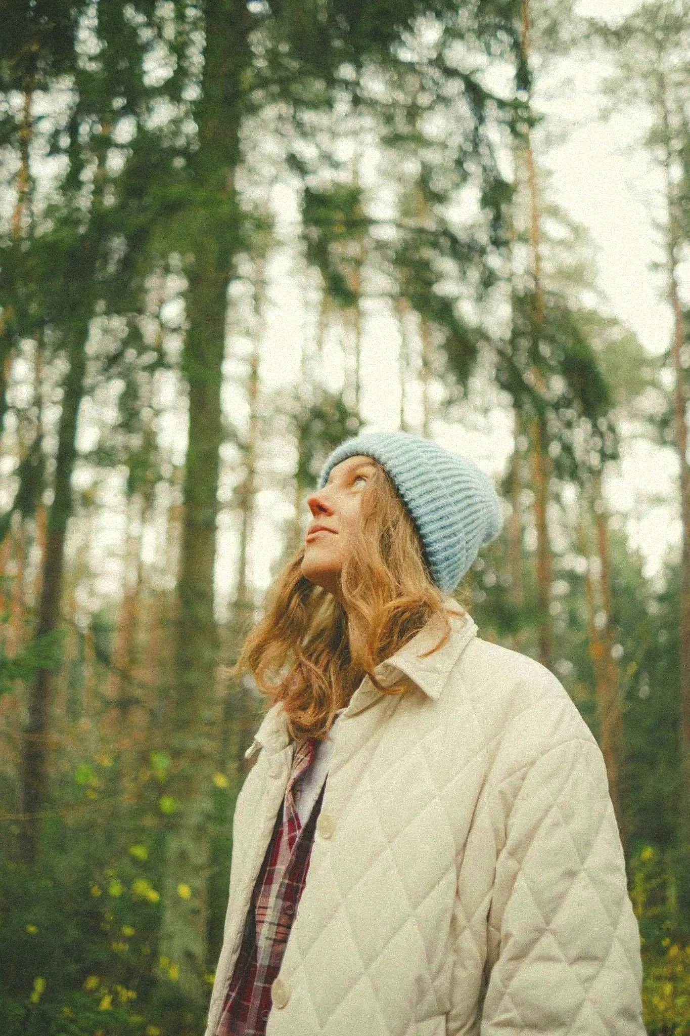 A young woman with curly red hair wearing a gray beanie and a cream-colored quilted jacket, standing outdoors after trauma therapy in a dense forest with tall, thin trees and green foliage in the background.