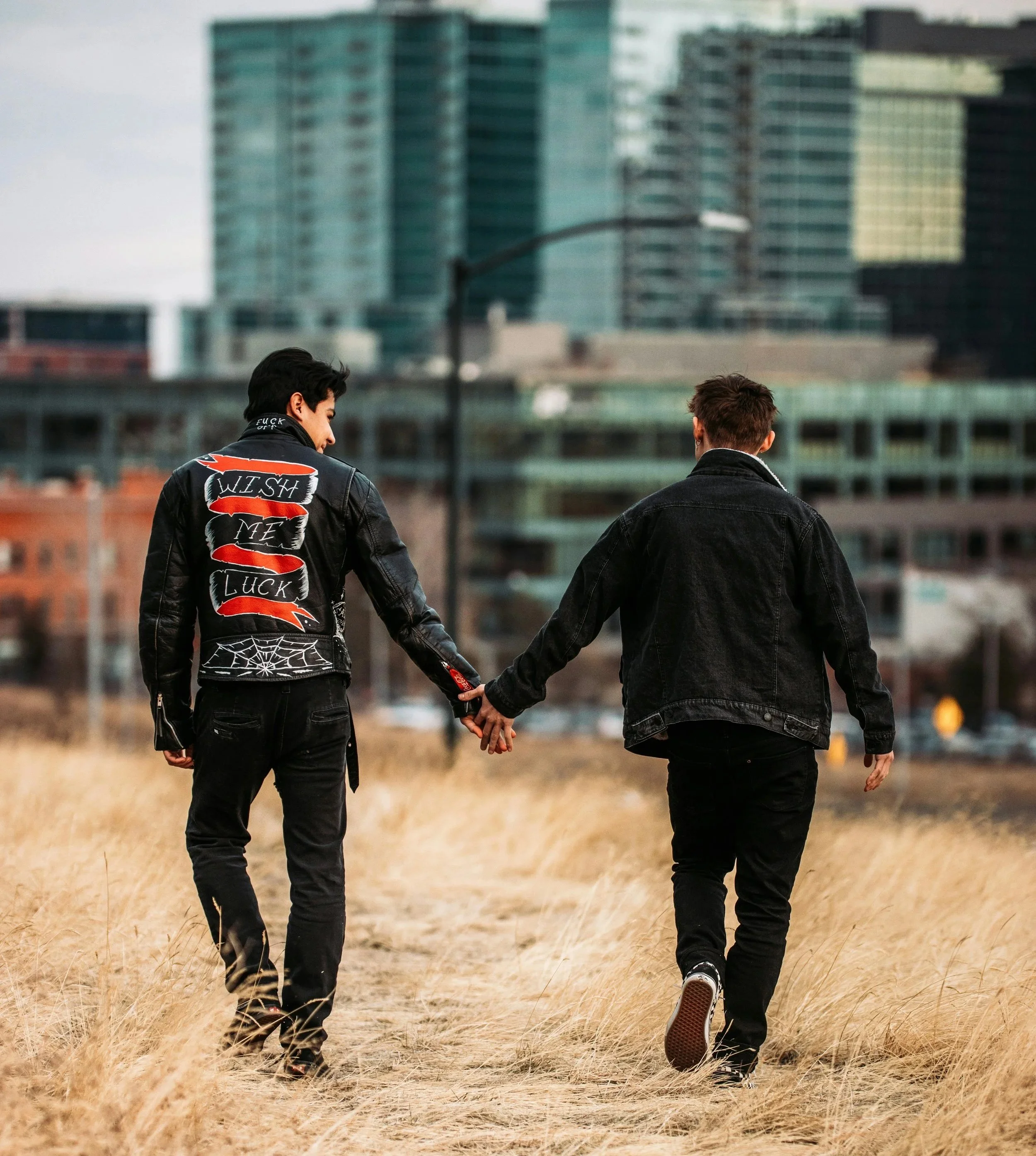 Two young men holding hands while walking through a field of tall, dry grass with a cityscape of modern high-rise buildings in the background.