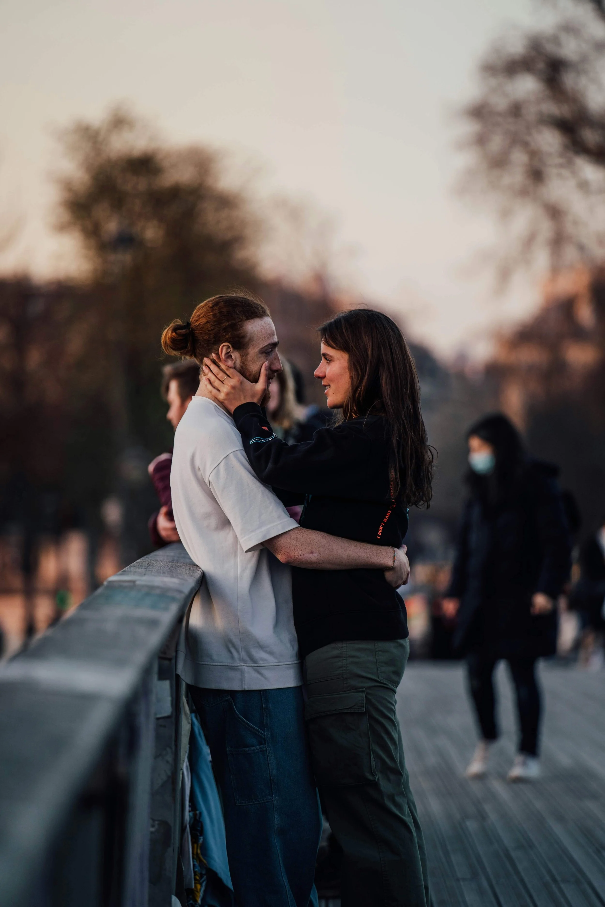 A young man and woman are standing close together on a bridge, gazing into each other's eyes. The man has red hair tied back in a bun, and the woman has long dark hair. The background shows other blurred people and trees during sunset.