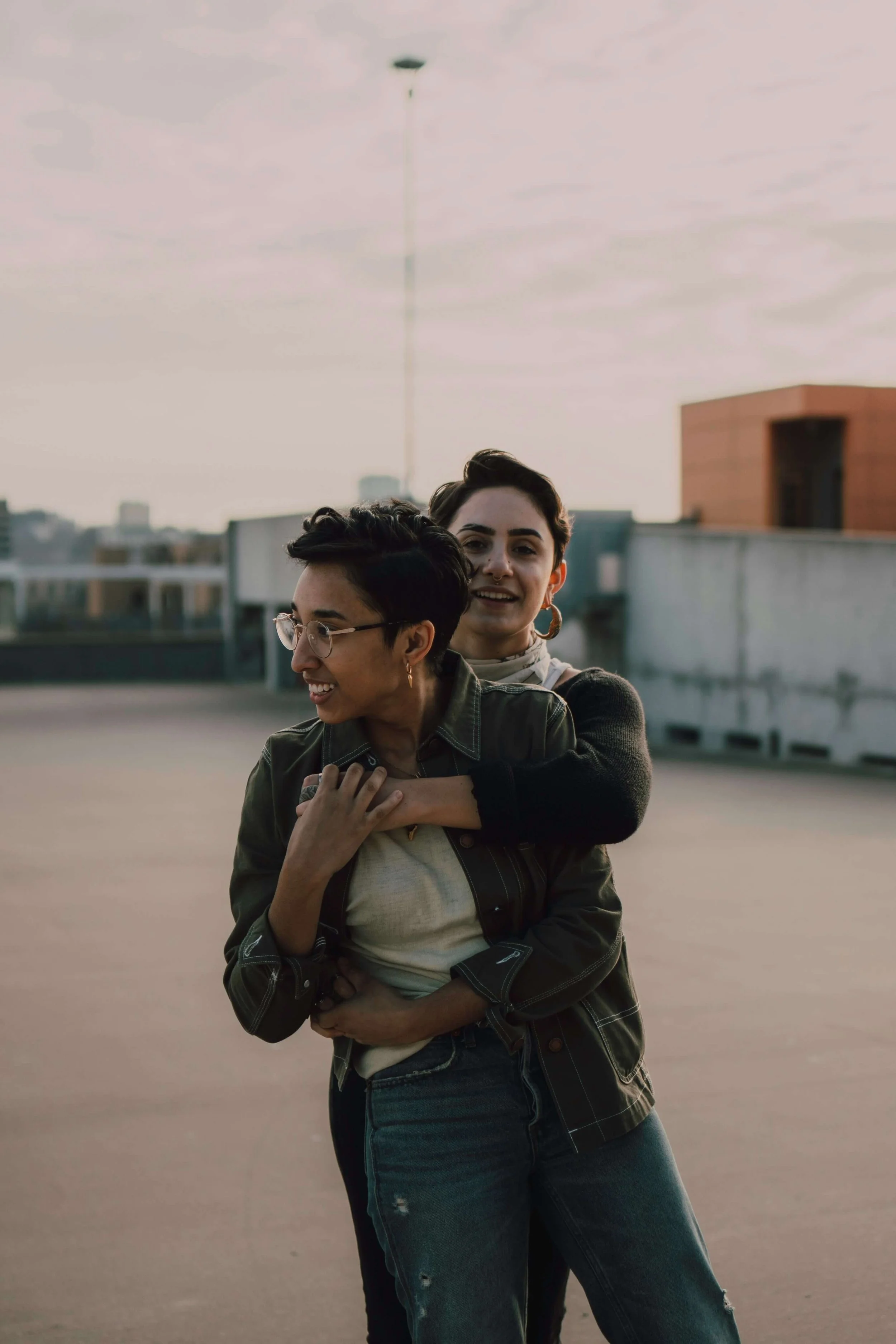 Two women standing closely together outdoors during daytime. One woman is wearing glasses and a green jacket, smiling and looking to the side. The other woman is behind her, with short dark hair, wearing earrings, and smiling at the camera. The background features a rooftop with buildings and an overcast sky.