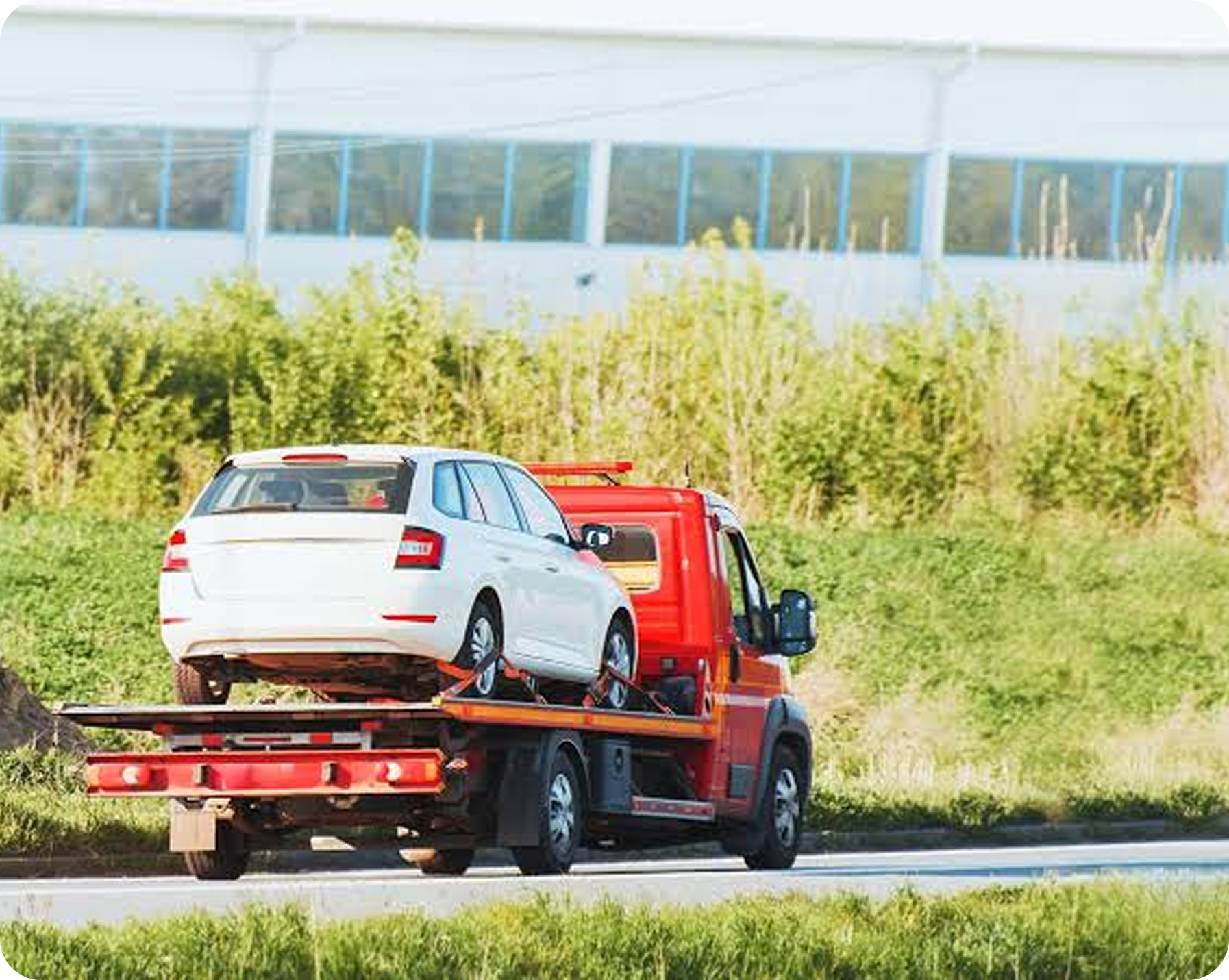 A white car loaded on a red tow truck driving on a highway with green grass and trees in the background.