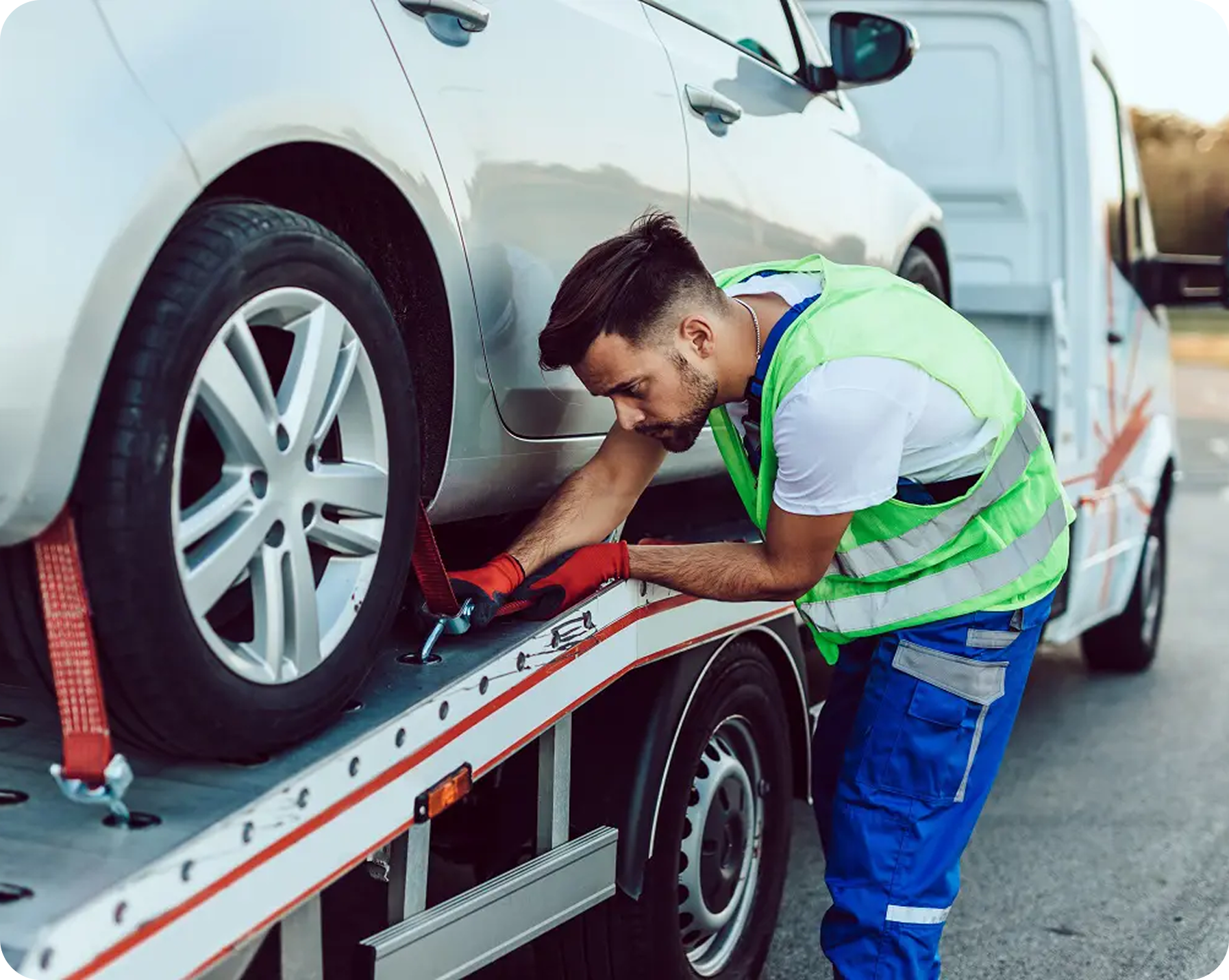 A man wearing a reflective safety vest and blue work pants inspecting a silver car on a flatbed tow truck, with the car secured with red straps.