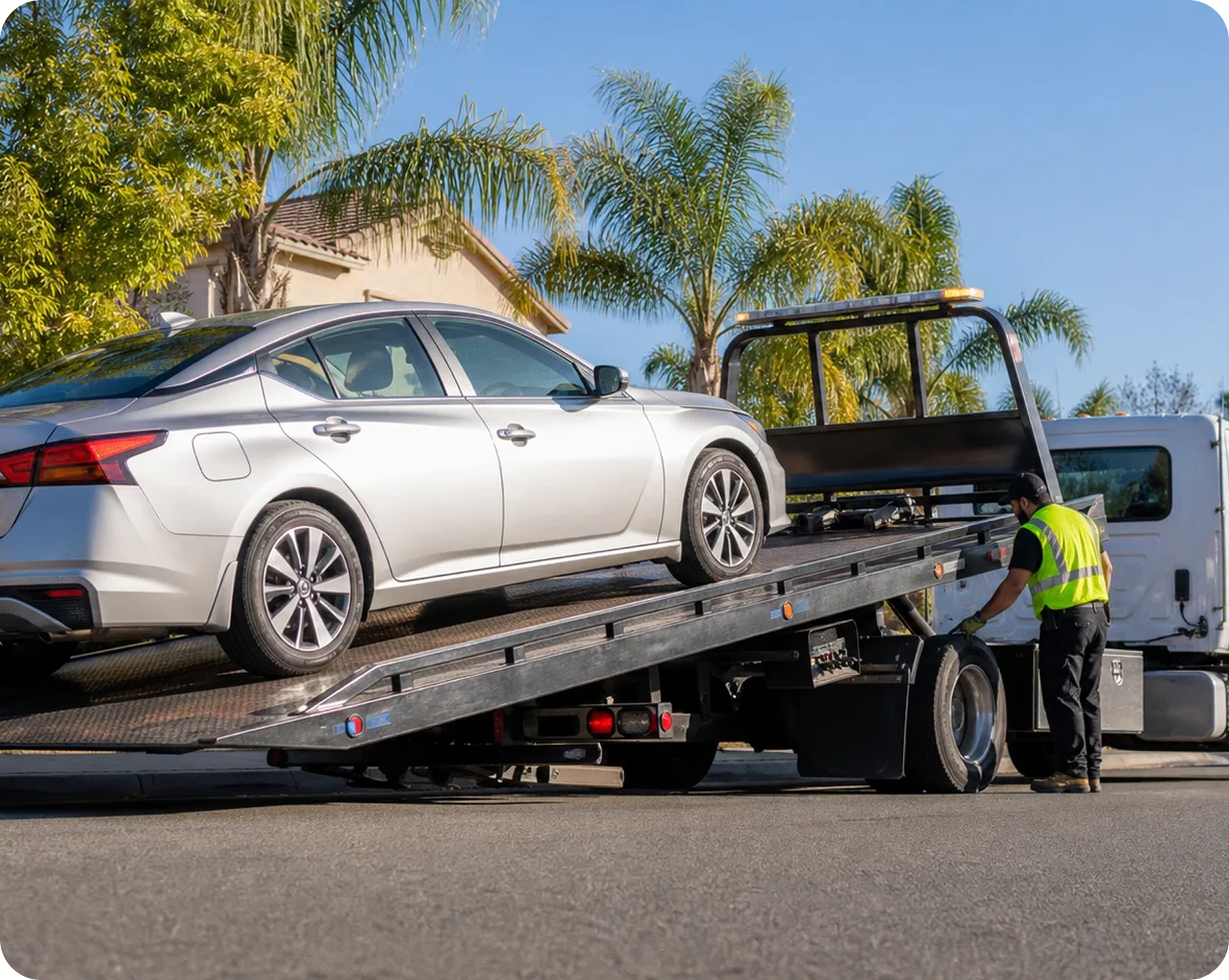 A white sedan being loaded onto a flatbed tow truck while a worker in a yellow safety vest assists. The background features palm trees and a house with a tiled roof under a clear blue sky.