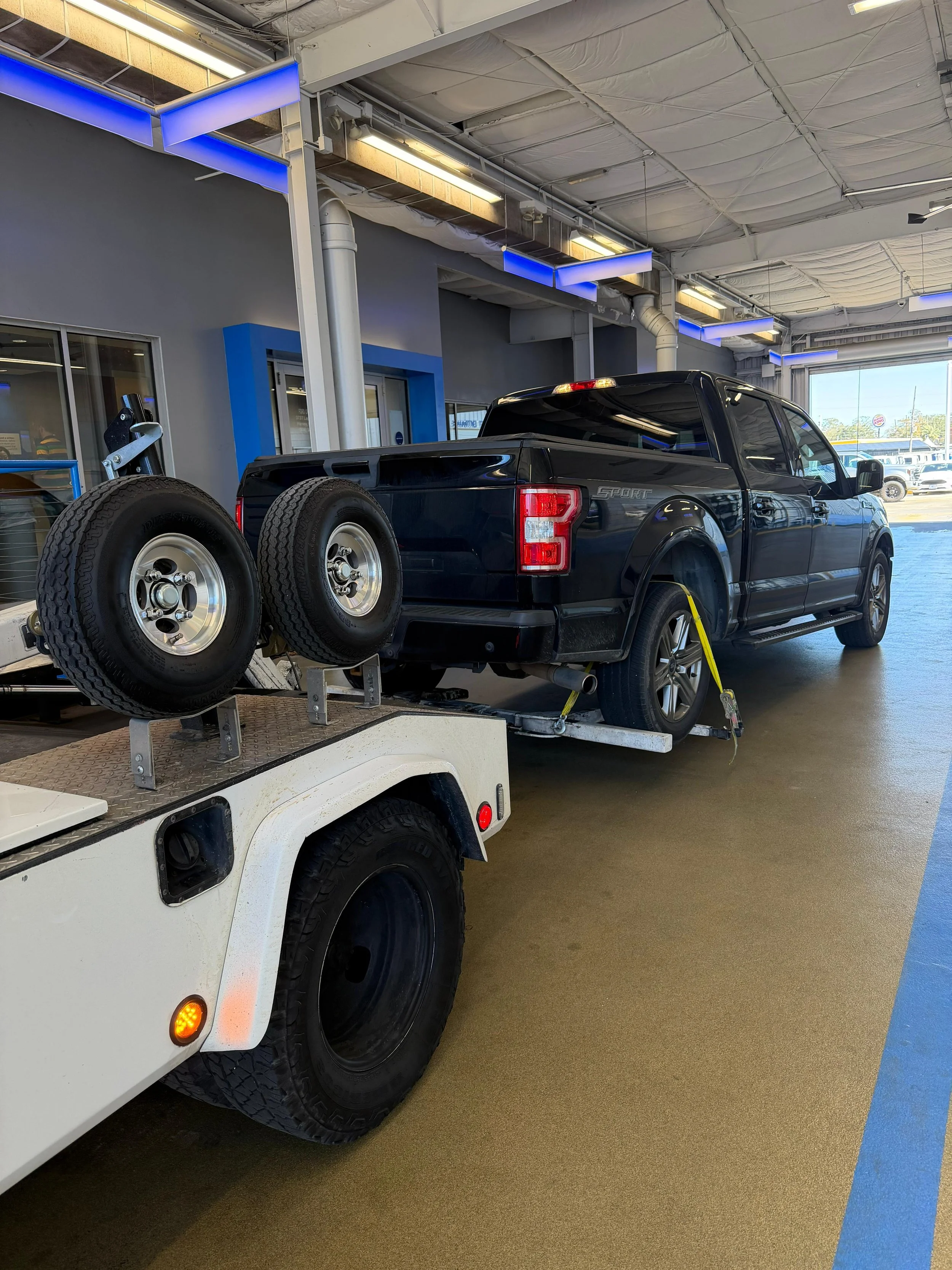 A black pickup truck on a flatbed tow truck with two spare tires mounted on its rear, inside a garage with gray walls and blue accents, bright ceiling lights, and a large open garage door showing cars and a parking lot outside.