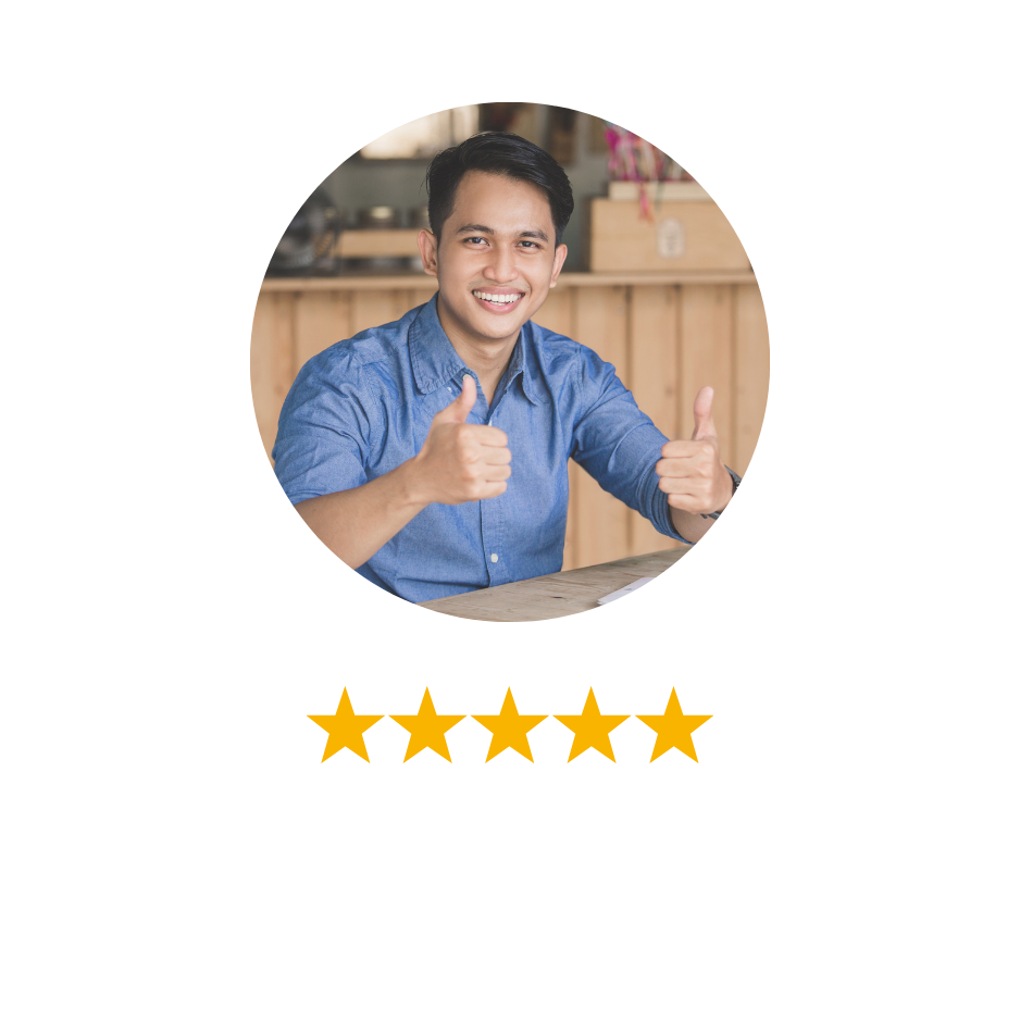 A young man in a blue shirt smiling and giving two thumbs up while sitting at a table in a cozy workspace.