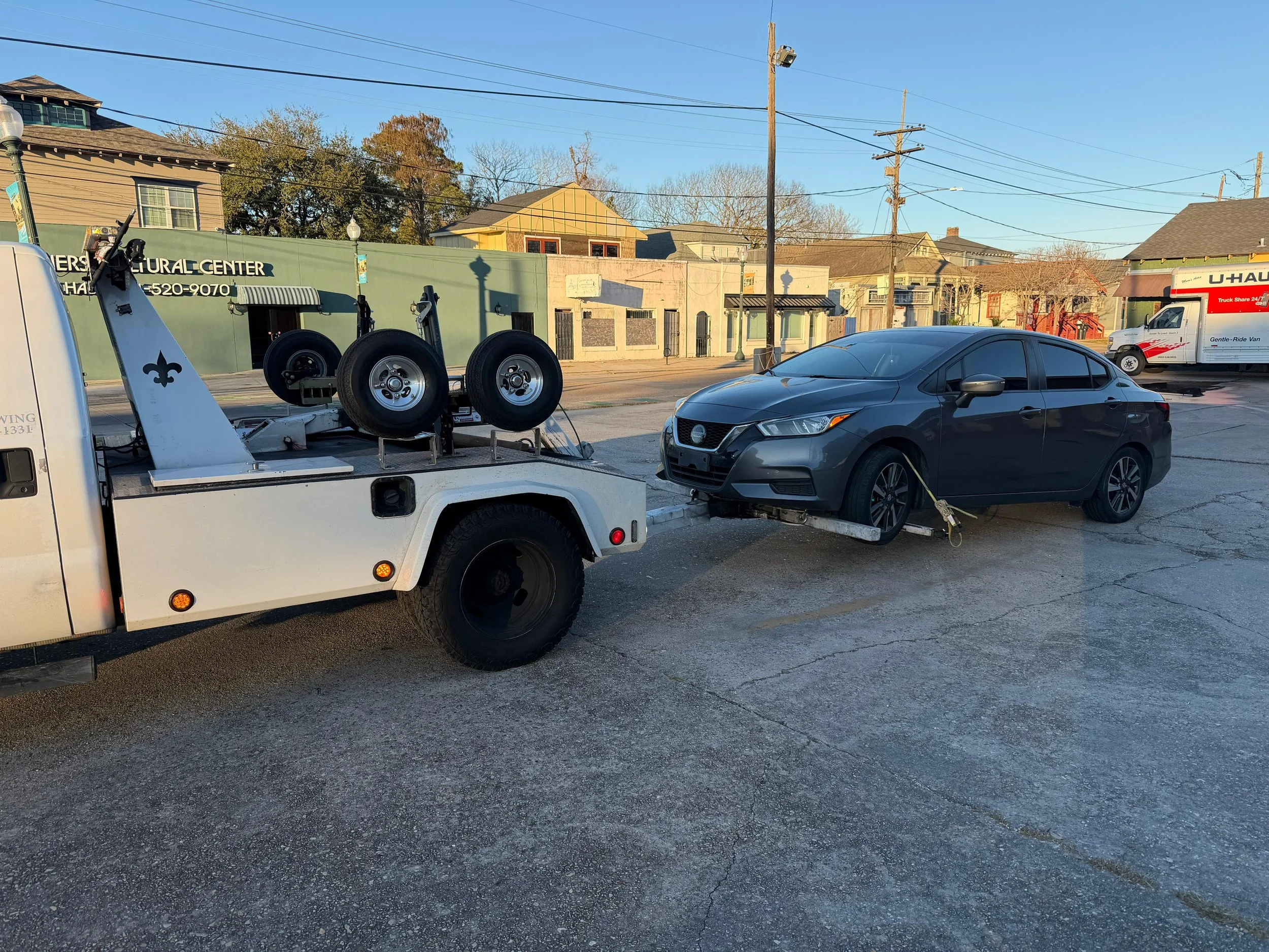 A parking lot with a black car on a flatbed towing trailer, some buildings in the background, utility poles and wires, a U-Haul truck, and clear blue sky.
