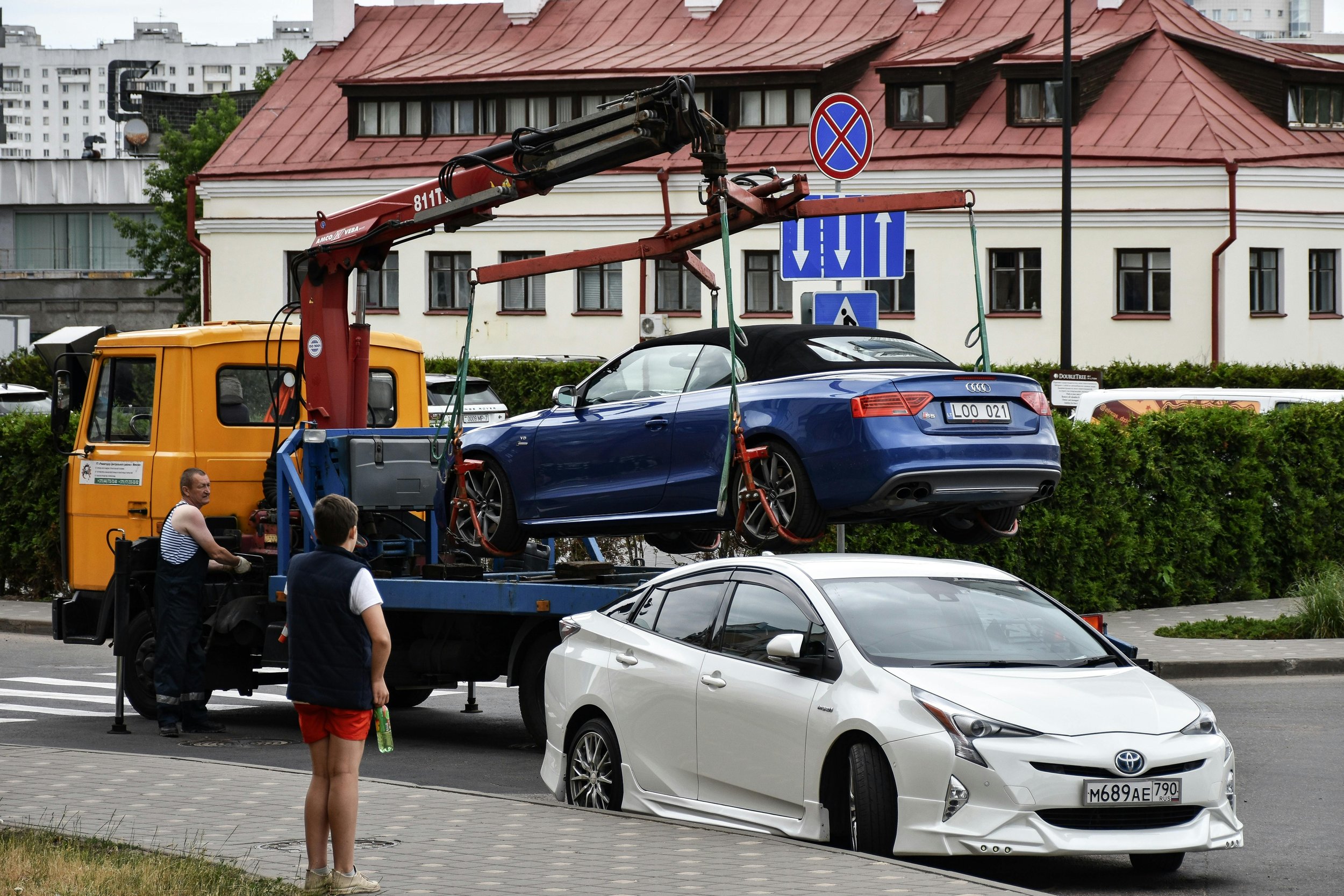A blue convertible car being lifted by a crane on a flatbed truck, with spectators nearby, in a city street with apartment buildings in the background.