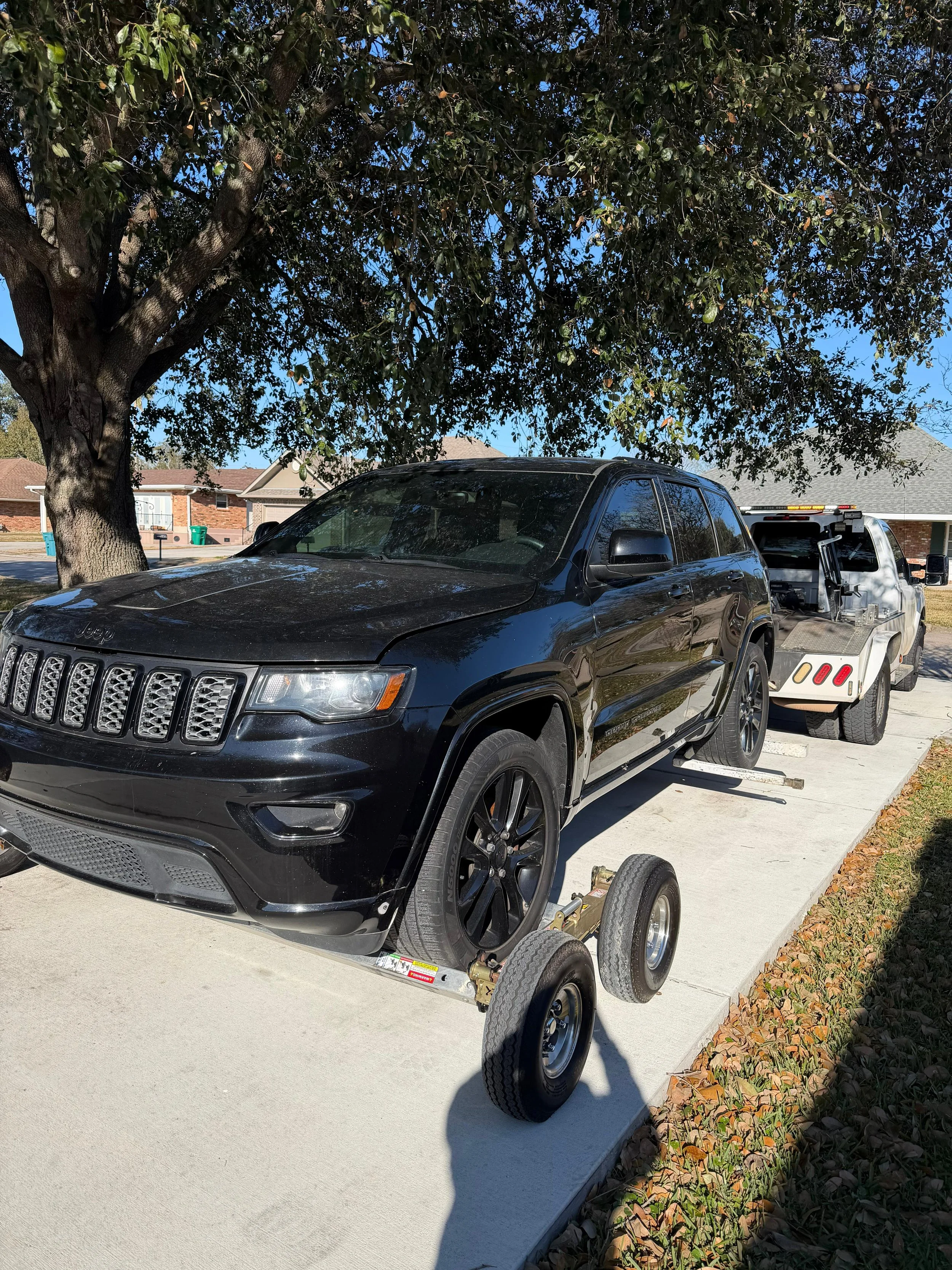 A black Jeep SUV is parked on a driveway next to a large tree with broad leaves. The SUV is attached to a small trailer with two wheels, which is parked on the same driveway.