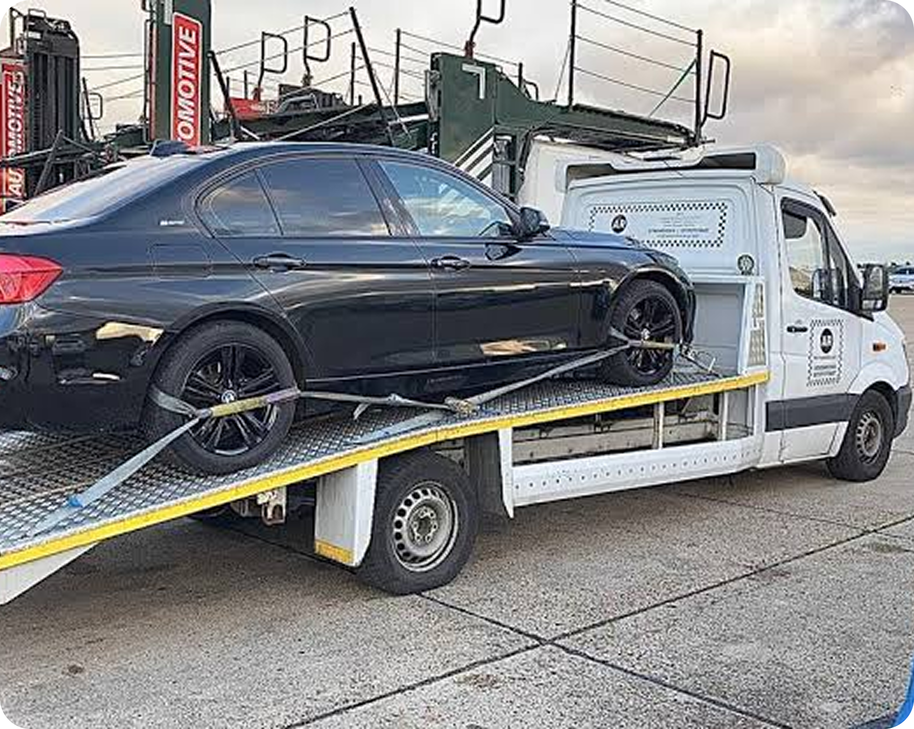 A black car on a flatbed tow truck, with a white vehicle behind it, possibly in a vehicle storage or transportation lot.