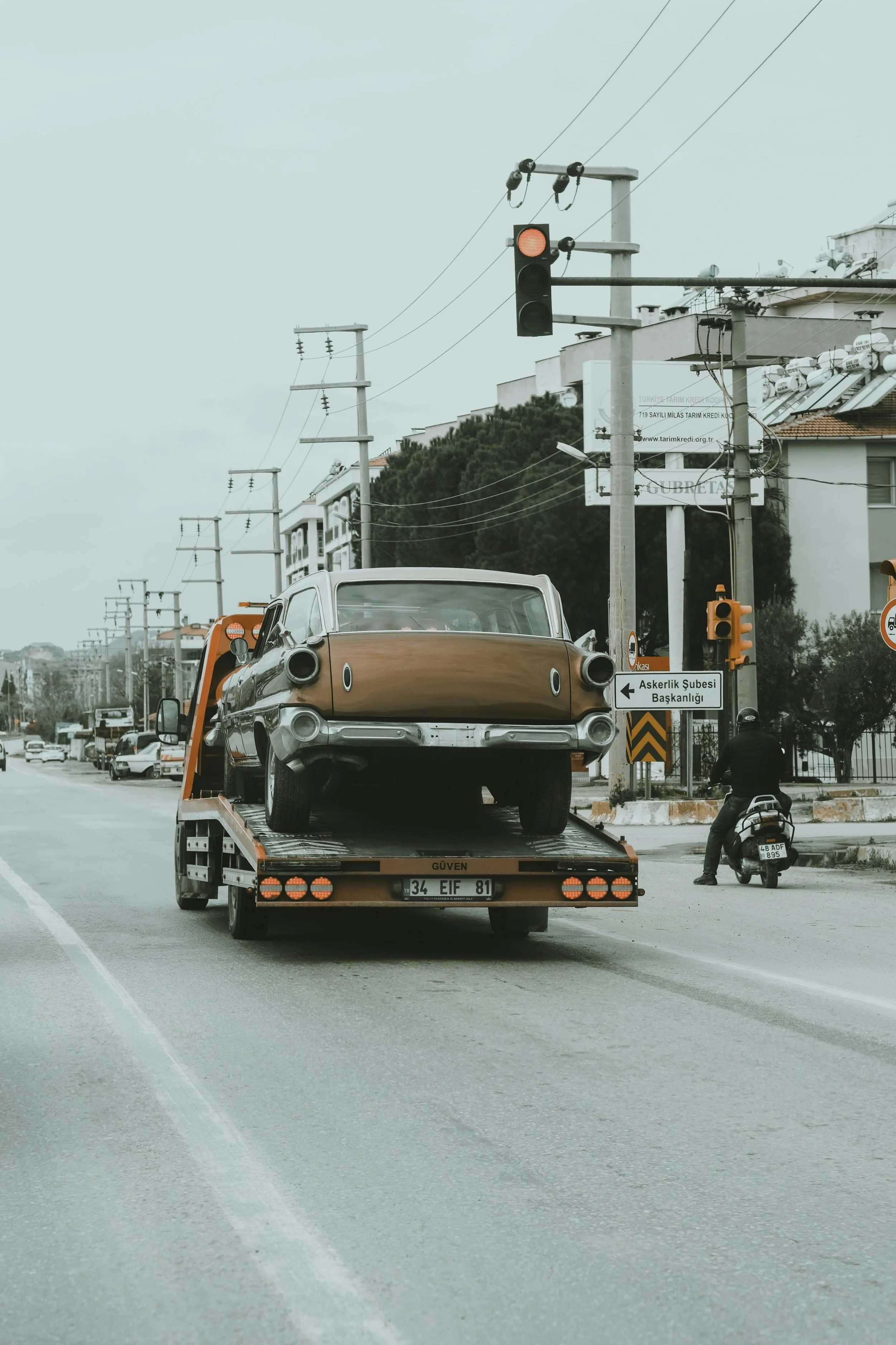 A vintage brown car on a flatbed tow truck on a city street, with traffic lights and electric poles in the background.