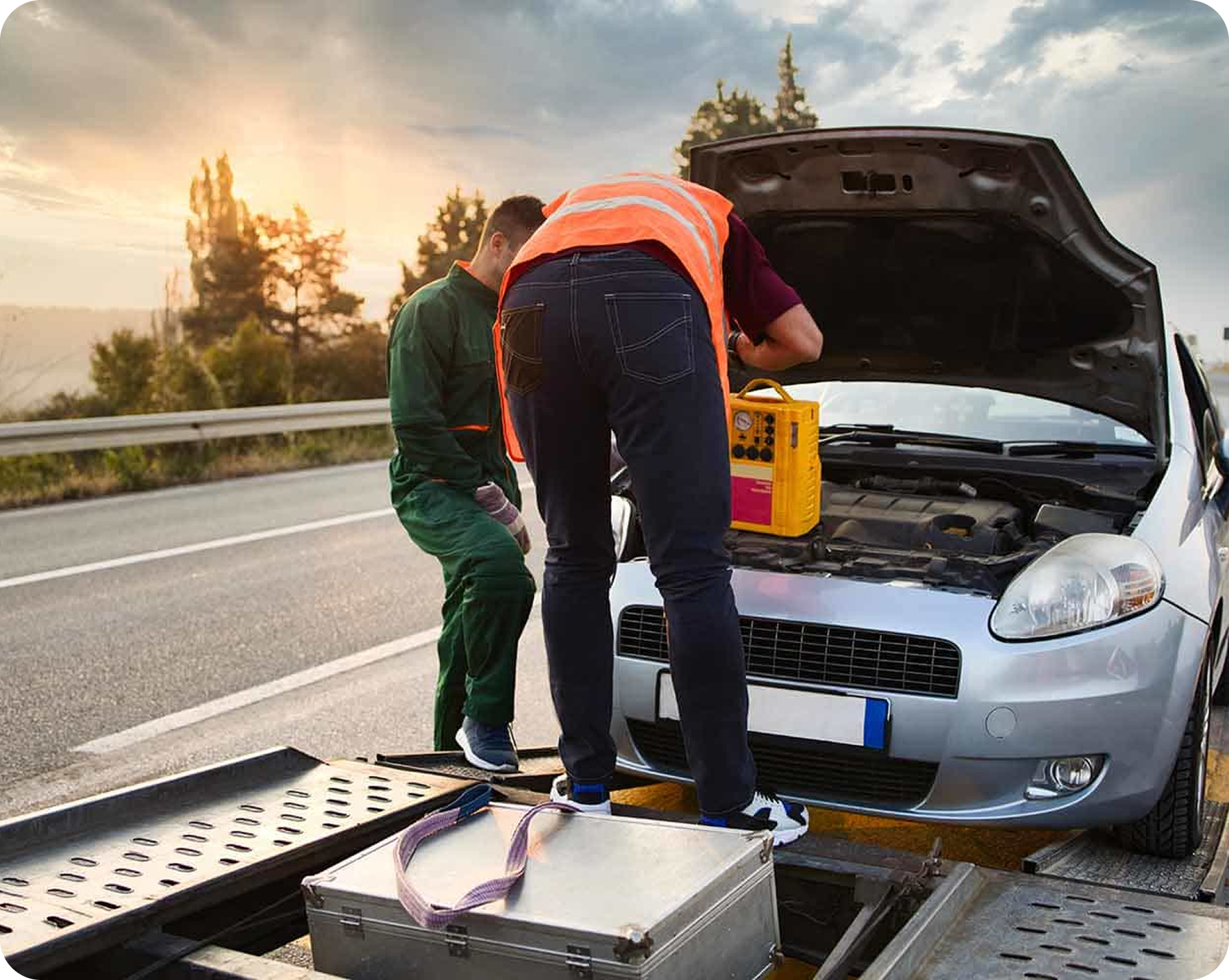 Two mechanics working on a white car with its hood open on the side of a road during sunset. One mechanic is wearing a green jumpsuit, and the other is wearing a maroon shirt with an orange safety vest. They are using a yellow diagnostic tool. There is a flatbed tow truck with a silver toolbox and a metallic ramp in the foreground.