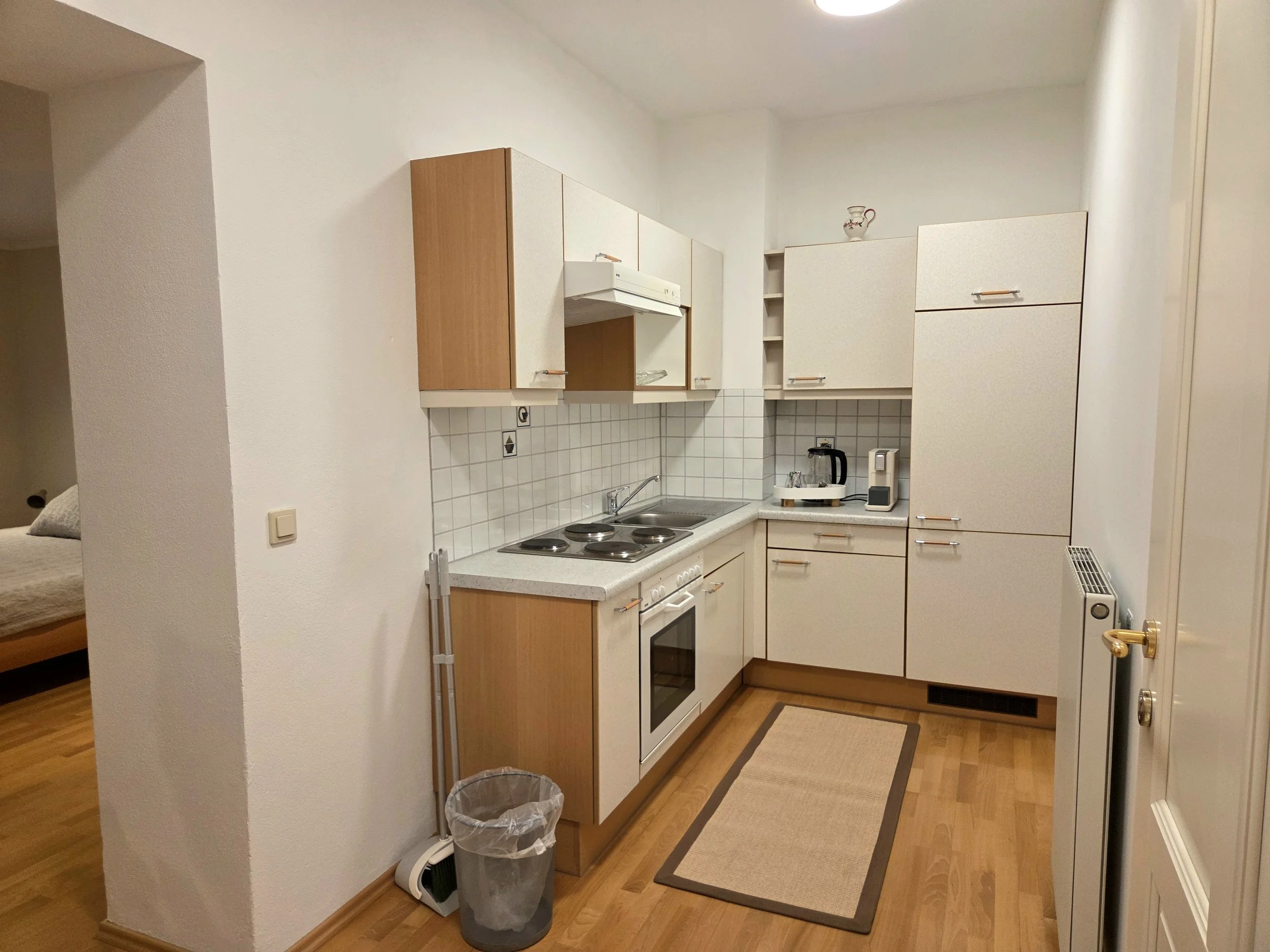 Kleine Küche mit weißen und Holzschränken, Herd, Spüle, Kaffeemaschine, Wasserkocher, im Hintergrund ein Schlafzimmer.