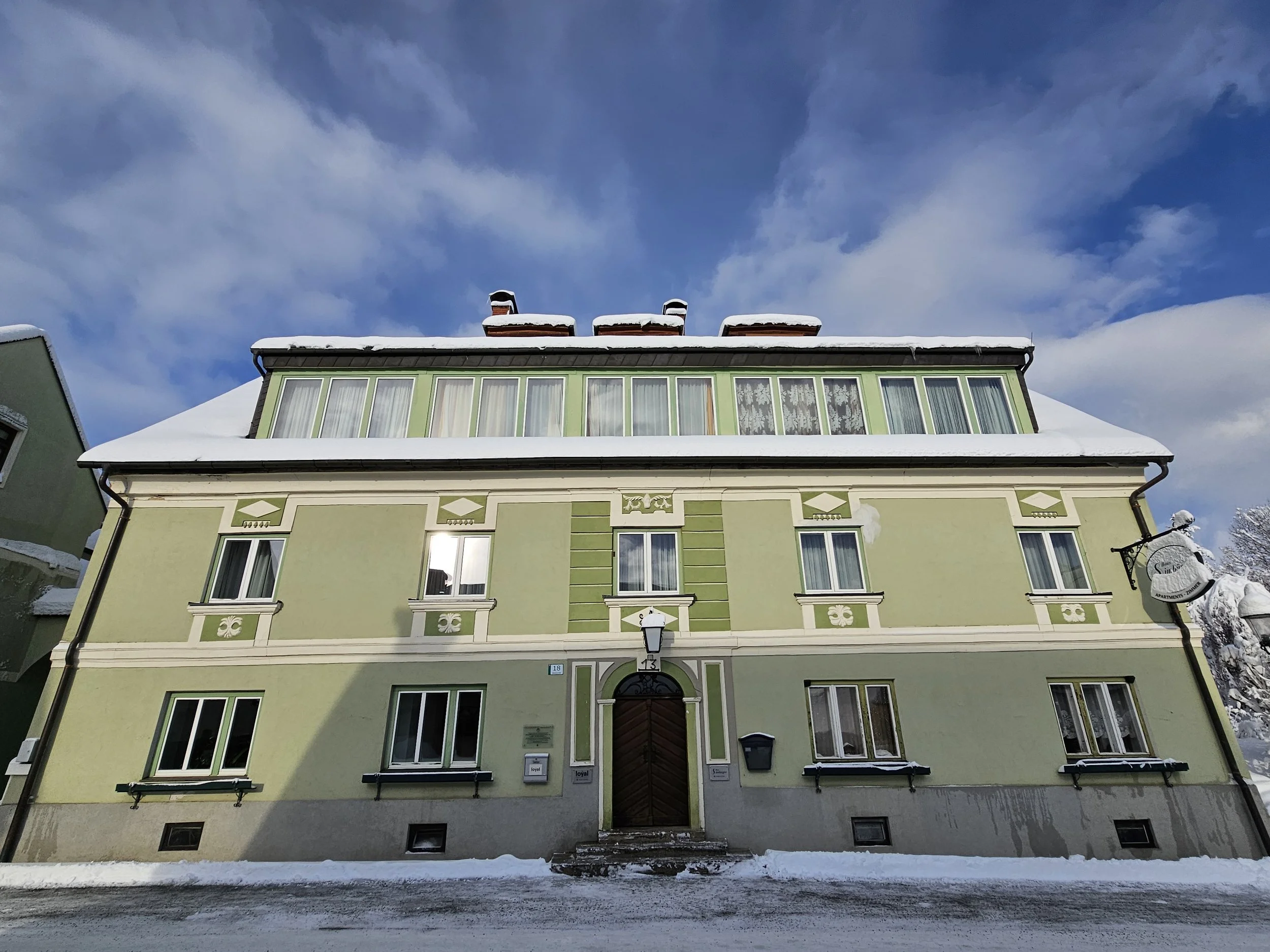 Ein mehrstöckiges, grünes Haus im winterlichen Schnee unter einem blauen Himmel mit Wolken.