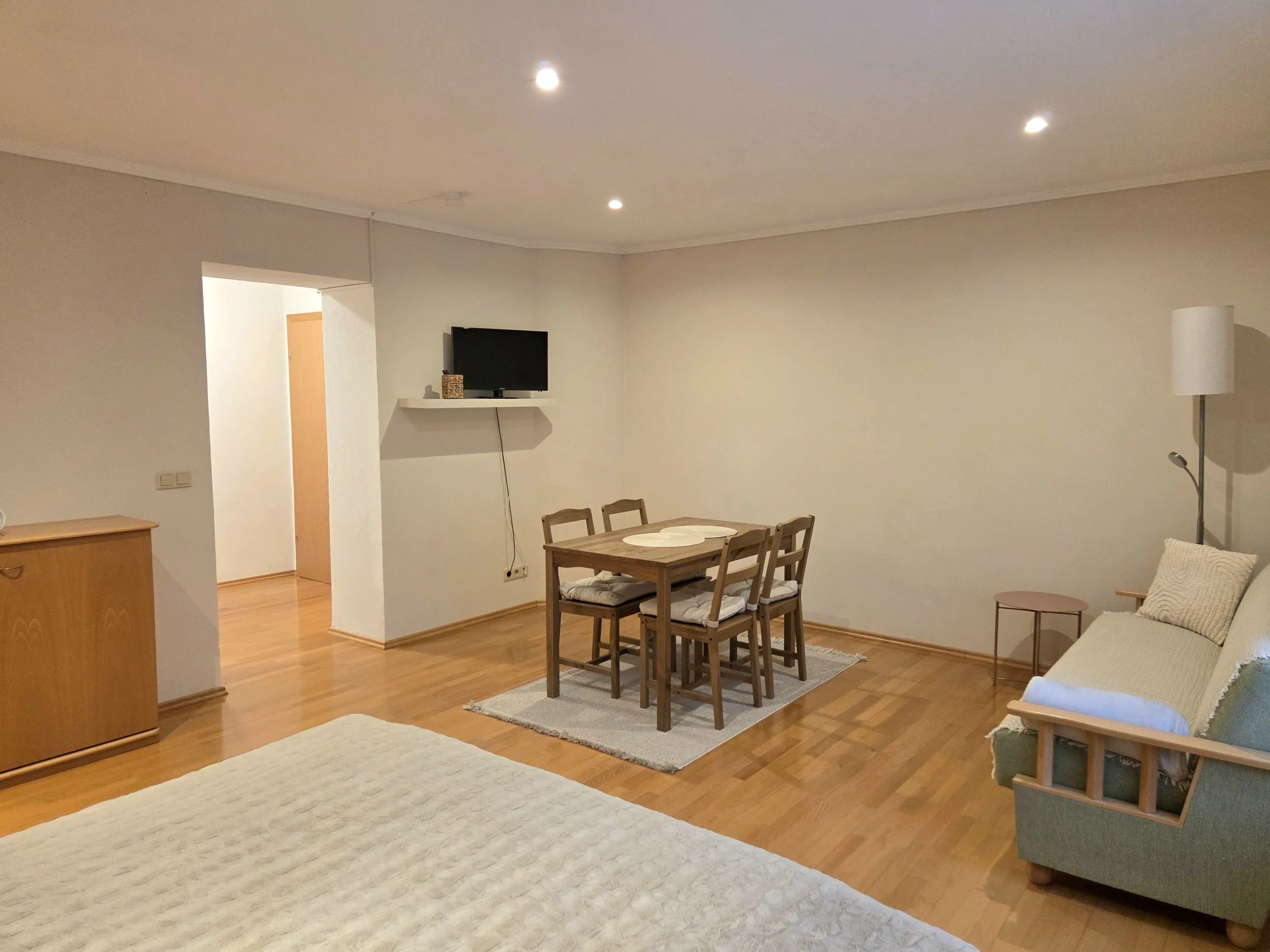 Innenansicht einer hellen, modernen Wohnung mit Holzfußboden, einem weißen Sofa, einem Holzstuhl und Esstisch, einem kleinen Fernseher an der Wand und Stehlampen.