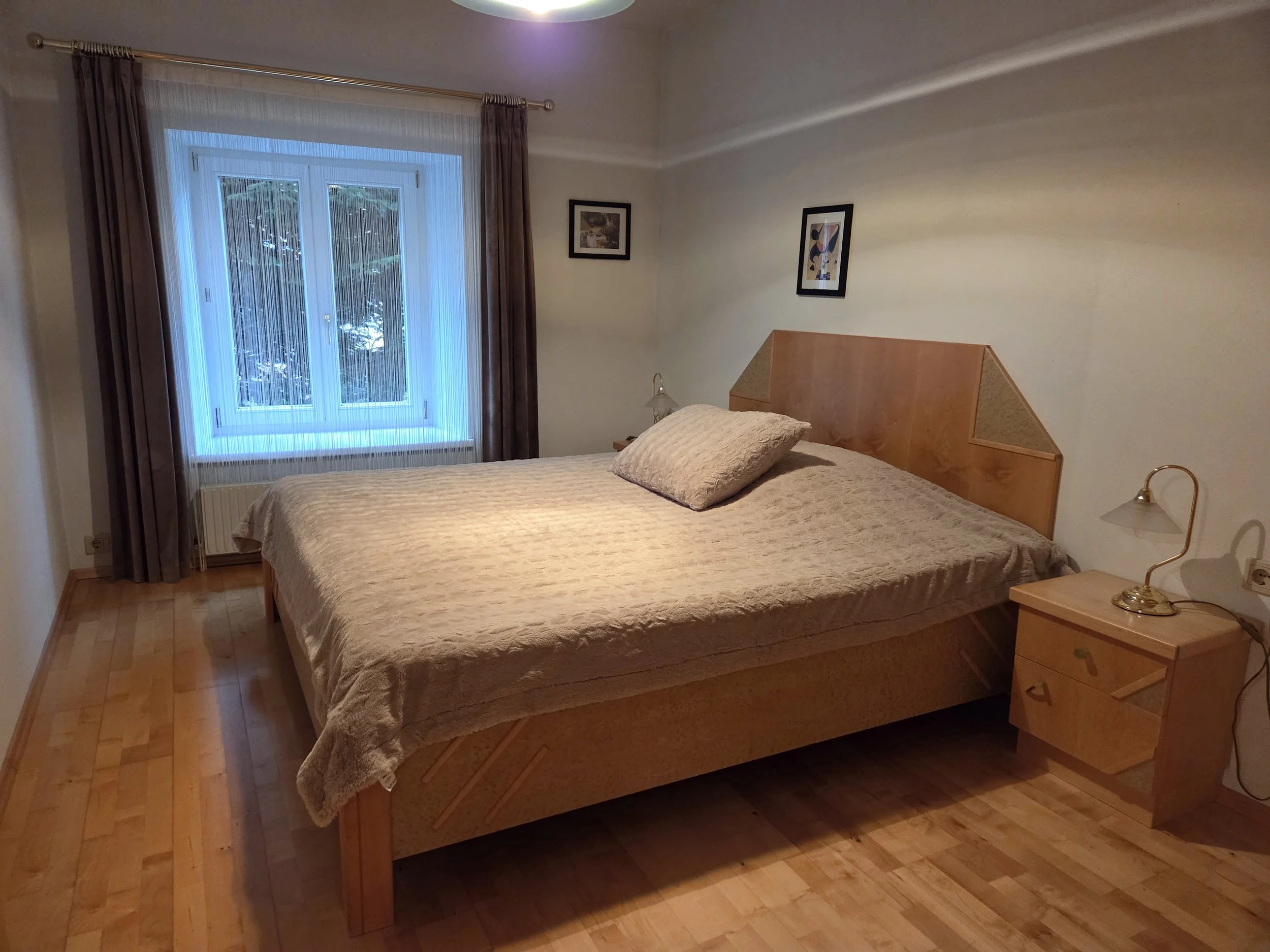 Schlafzimmer mit Fenster, Holzboden, Doppelbett, Nachttischlampen und Bildern an der Wand.