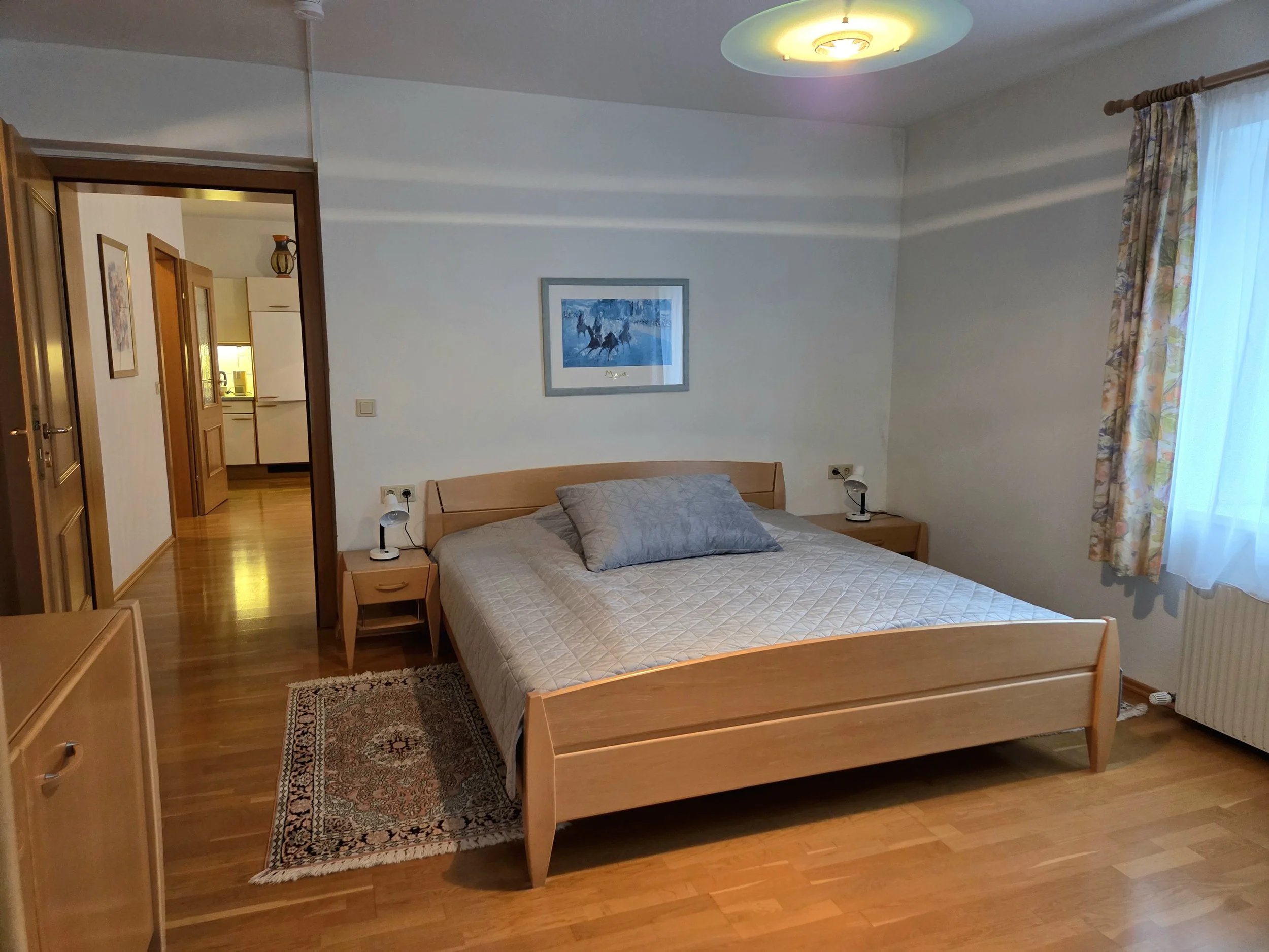 Schlafzimmer mit Doppelbett, zwei Nachttischen mit Lampen, Kunstwerk an der Wand, Gardinen am Fenster, Holzboden, Deckenleuchte.