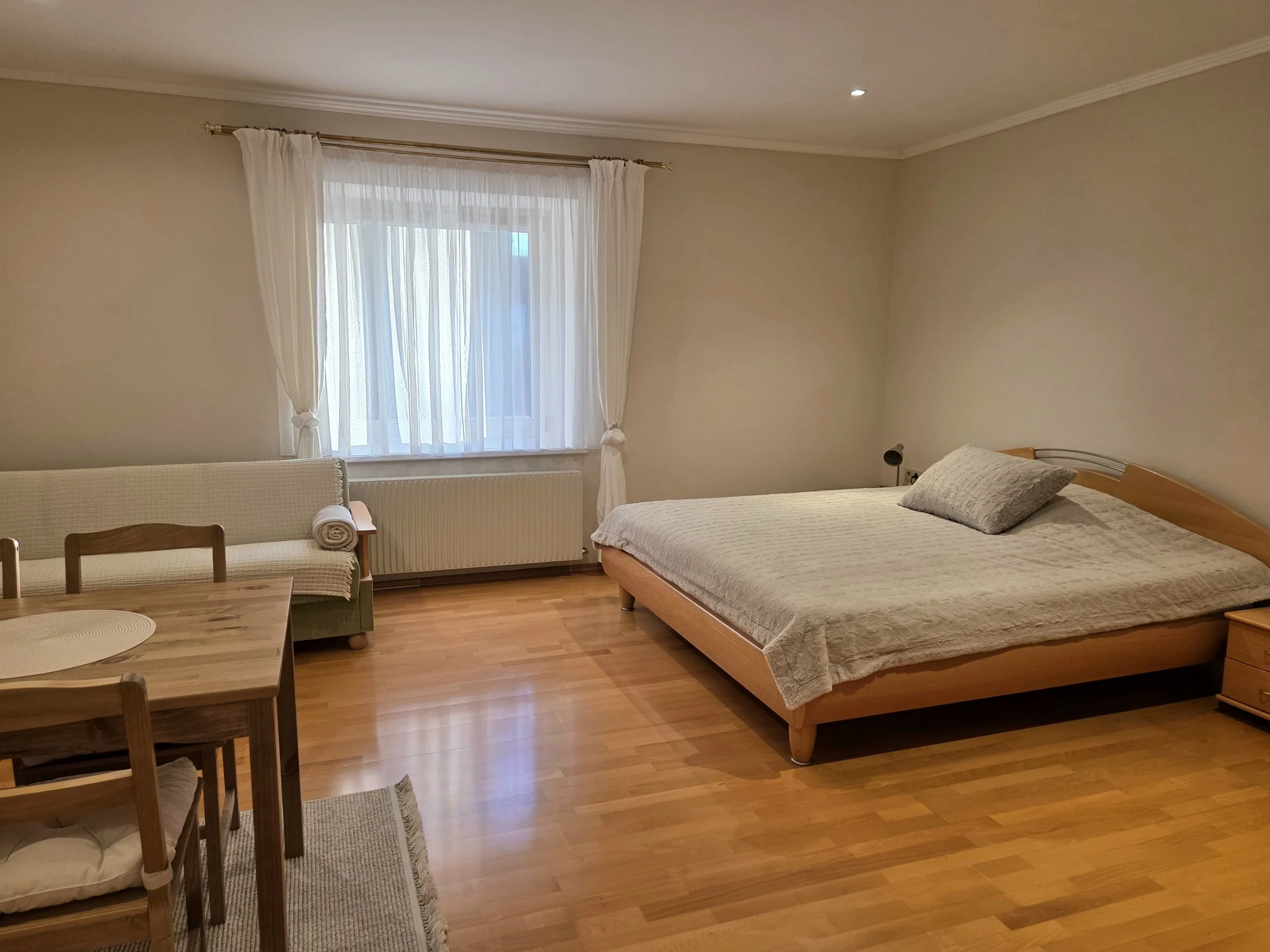 Schlafzimmer mit Holzfußboden, einem großen Bett mit grauem Bettzeug, einem Nachttisch, einem Sofa, einem Esstisch mit Stühlen, einer Fenster mit weißen Vorhängen und einem Heizkörper darunter.