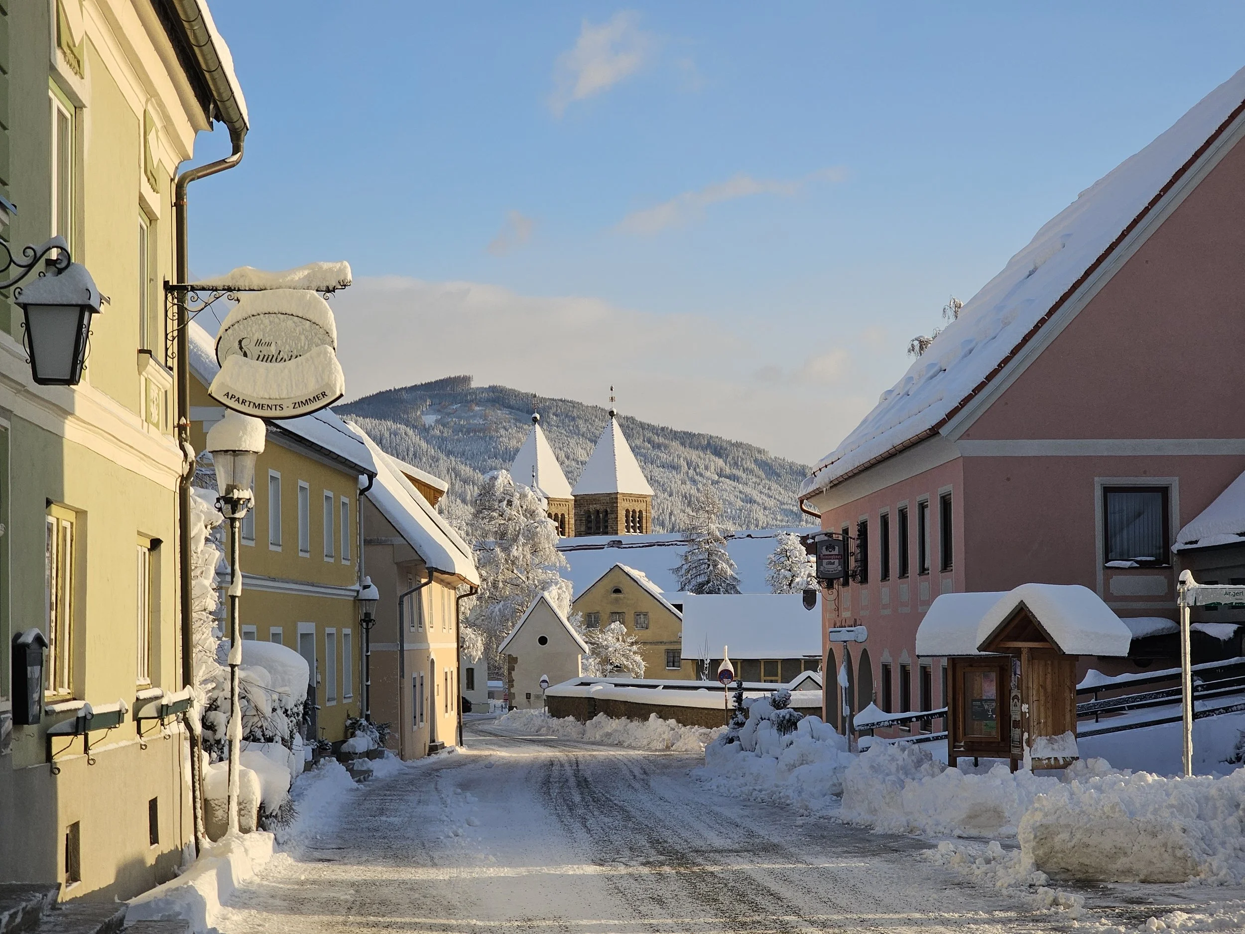 Eine verschneite Straße in einem europäischen Dorf, umgeben von bunten Häusern mit Schnee bedeckten Dächern und einem Berg im Hintergrund.