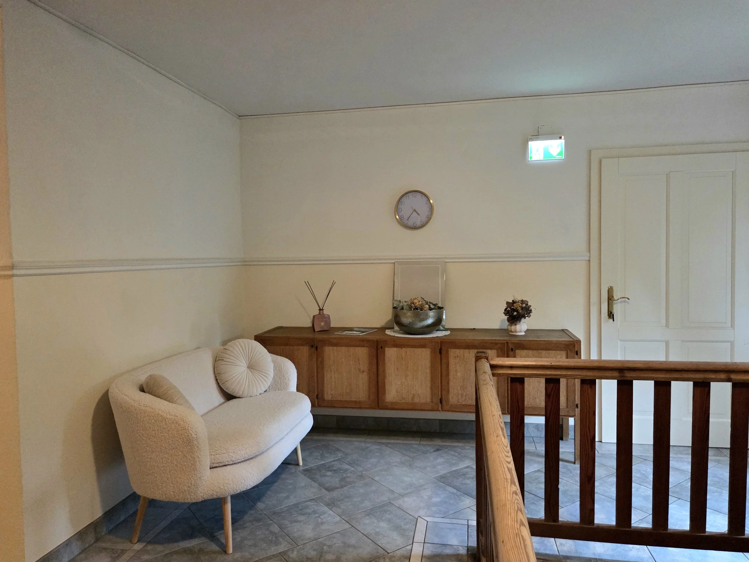Ein Wartebereich mit einer weißen gemütlichen Couch, einem Holzsideboard mit Dekorationsgegenständen, einer Standleuchte und einer Uhr an der Wand, Fliesenboden.