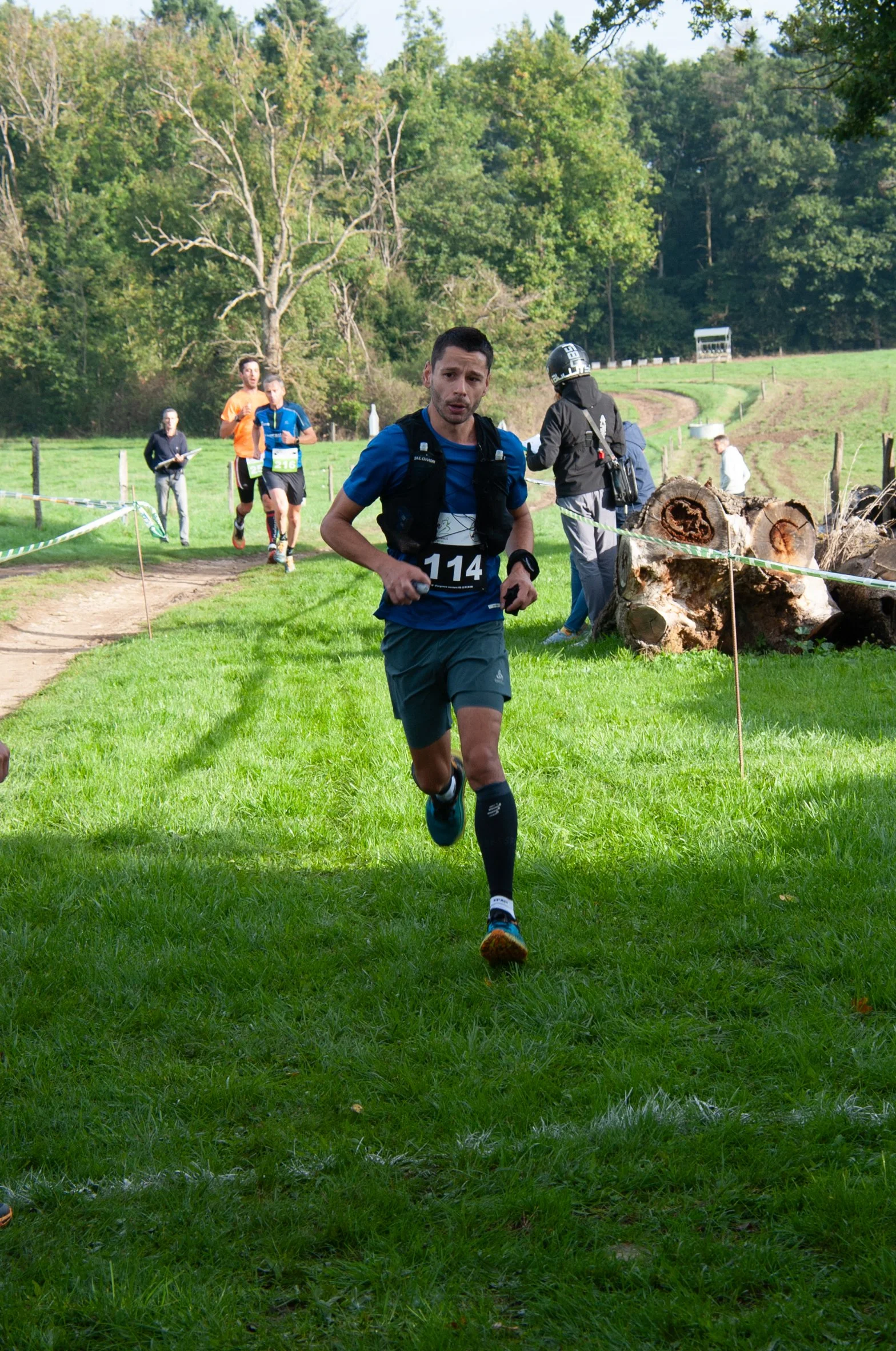 Un homme court lors d'une course en plein air dans un champ vert avec des arbres en arrière-plan, portant un t-shirt bleu, un short gris, des chaussettes hautes noires, un entraîneur ou spectateur à côté, et d'autres coureurs derrière.