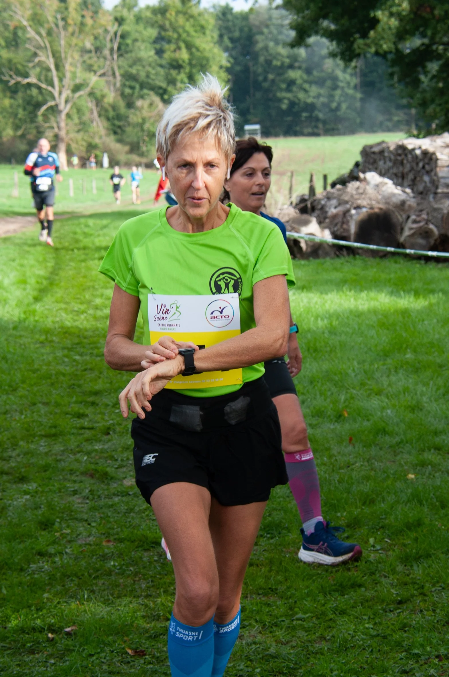 Une femme en tenue de course à pied portant un dossard, en pleine course dans un environnement naturel verdoyant.