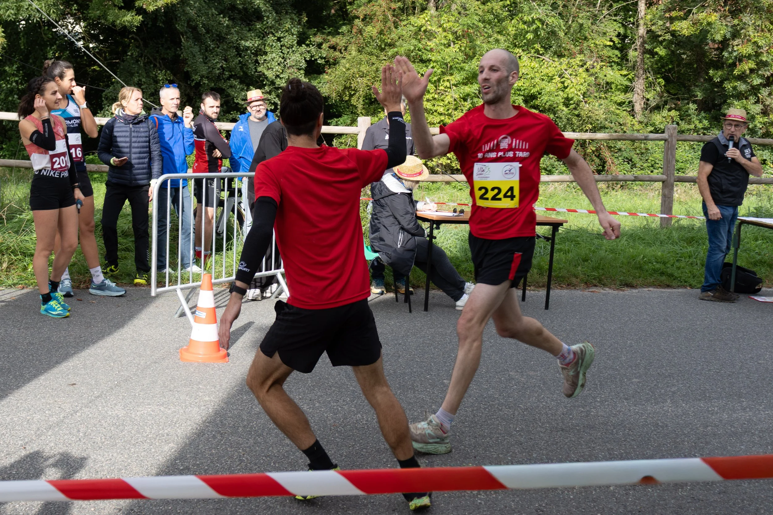 Deux coureurs franchissent la ligne d'arrivée en haut d'une course, se saluant ou se félicitant. Un groupe de spectateurs et officiels les regarde, avec des arbres en arrière-plan.