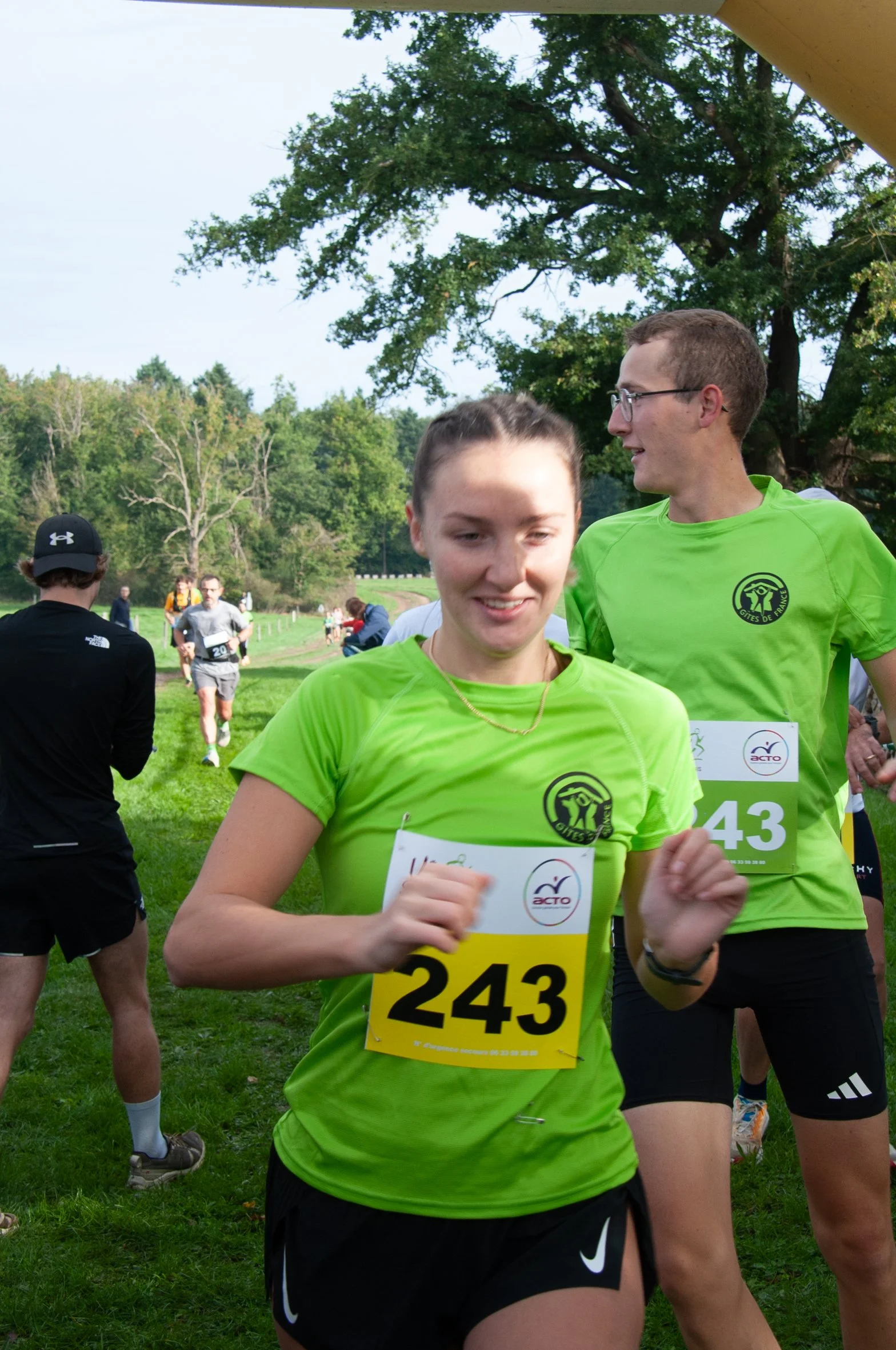 Deux coureurs portant des maillots verts lors d'une course, en train de s'échauffer ou de finir une étape, avec un groupe de participants en arrière-plan dans un parc vert avec des arbres.