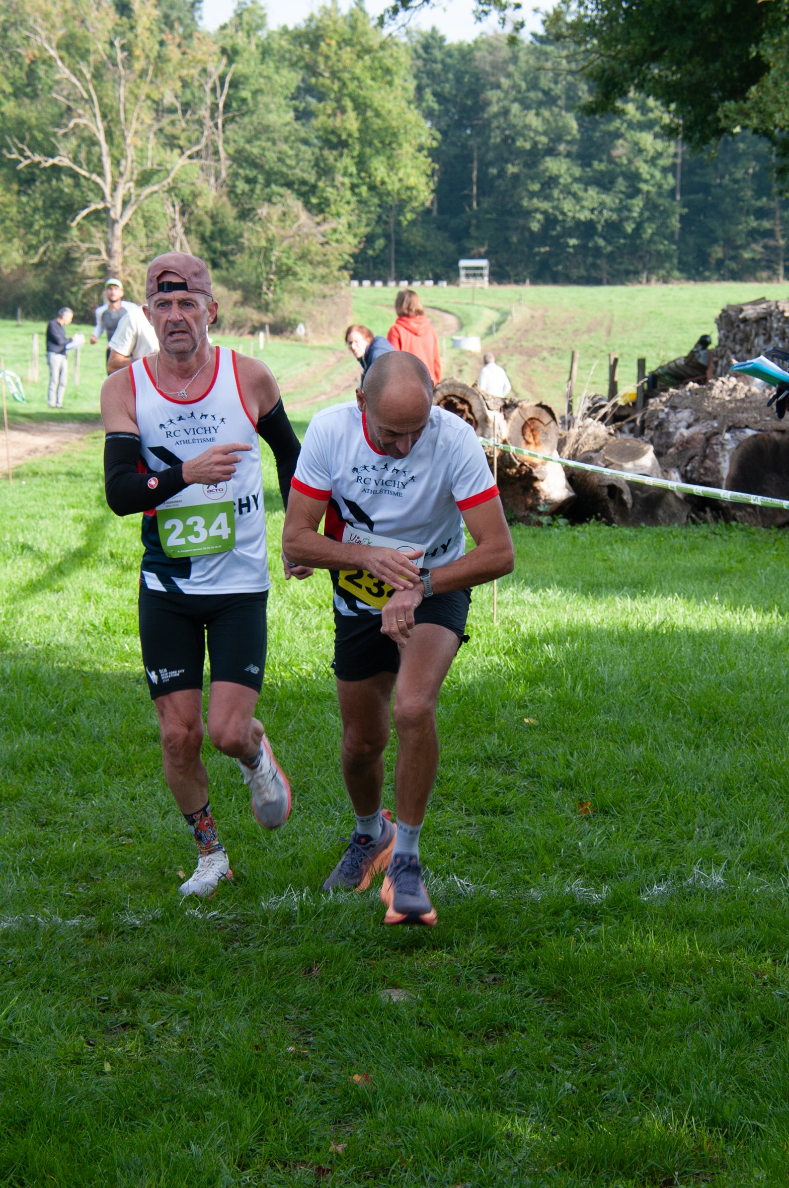 Deux hommes en course dans un parc vert lors d'une compétition, portant des maillots blancs avec des numéros 234 et 235.
