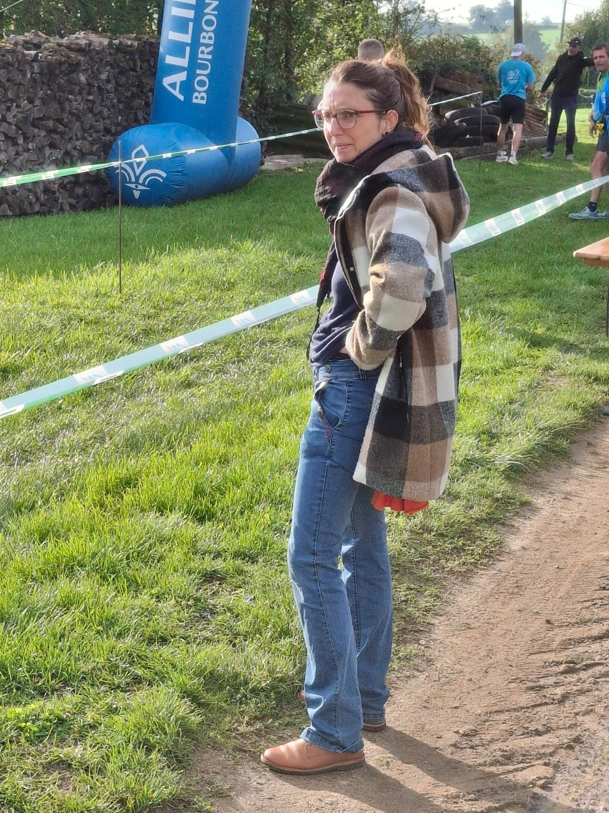 Femme portant une veste à carreaux, jeans et lunettes, se tenant sur un sentier herbeux à côté d'une corde de délimitation, lors d'un événement en plein air.