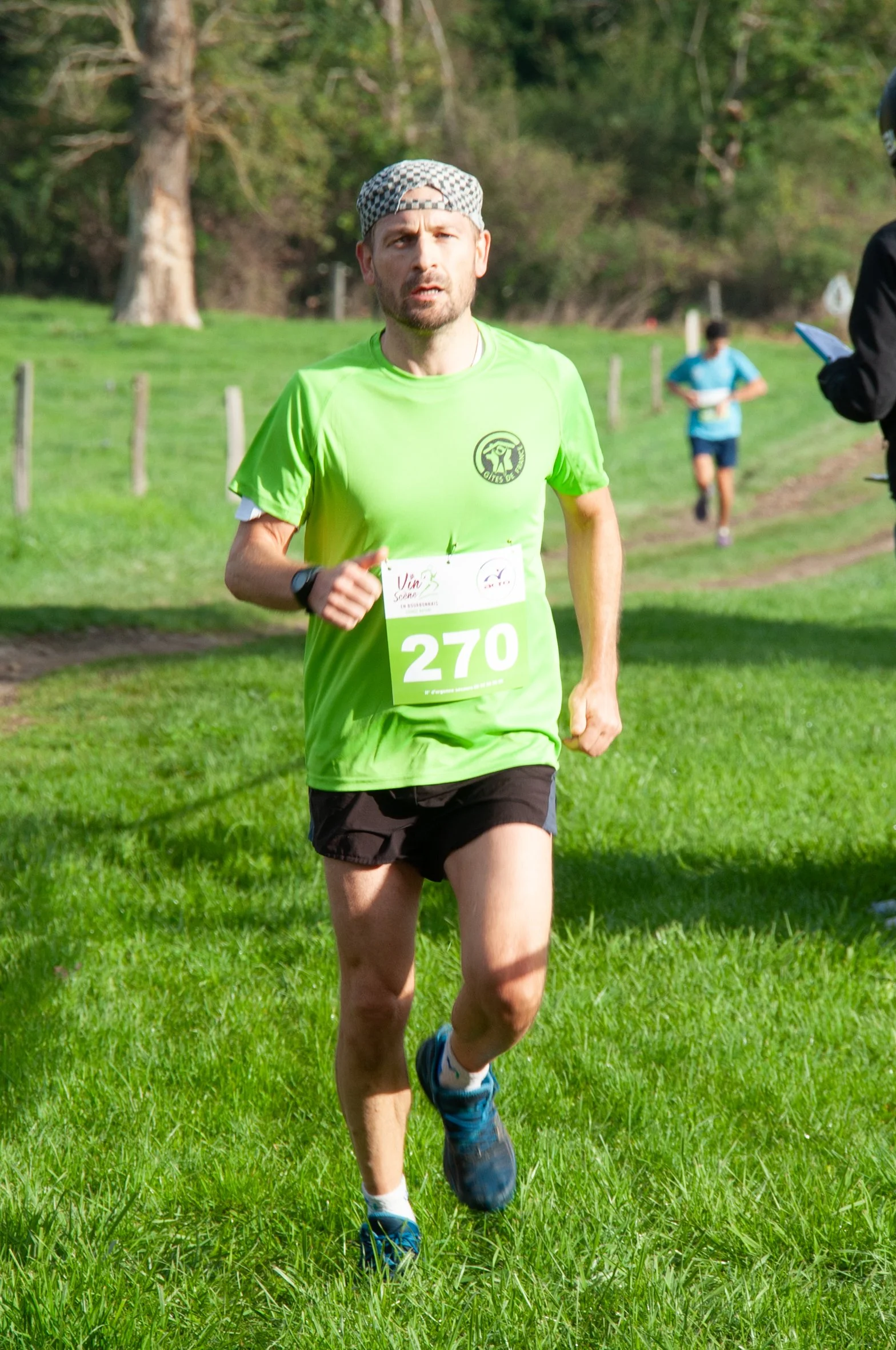 Un homme court lors d'une course en plein air, portant un t-shirt jaune fluo avec le numéro 270, un short noir, une montre et un bonnet, sur un fond de nature verte.