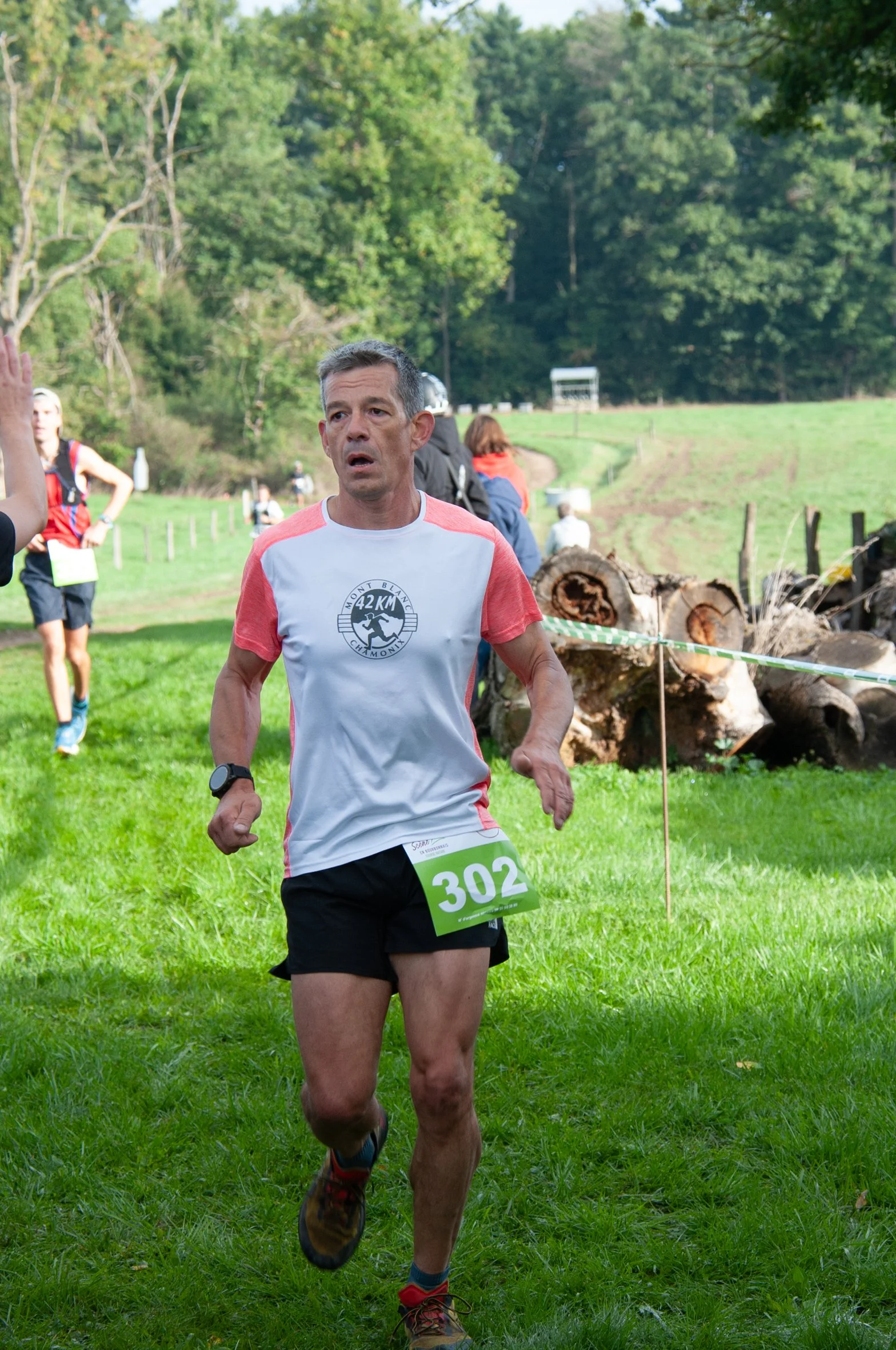 Un homme participe à une course en plein air dans un environnement naturel, portant un T-shirt blanc et rayé rouge, un short noir, et une montre connectée. Il porte un dossard numéro 302. Des arbres et un tronc d'arbre coupé sont visibles en arrière-