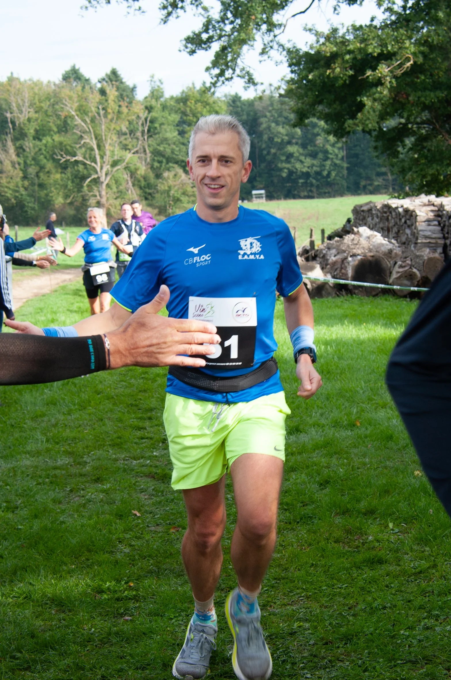 Un homme court lors d'une course en plein air, portant un t-shirt bleu, un short jaune fluorescent et des chaussures de sport, accueillant des encouragements. En arrière-plan, d'autres coureurs et un paysage verdoyant avec des arbres.