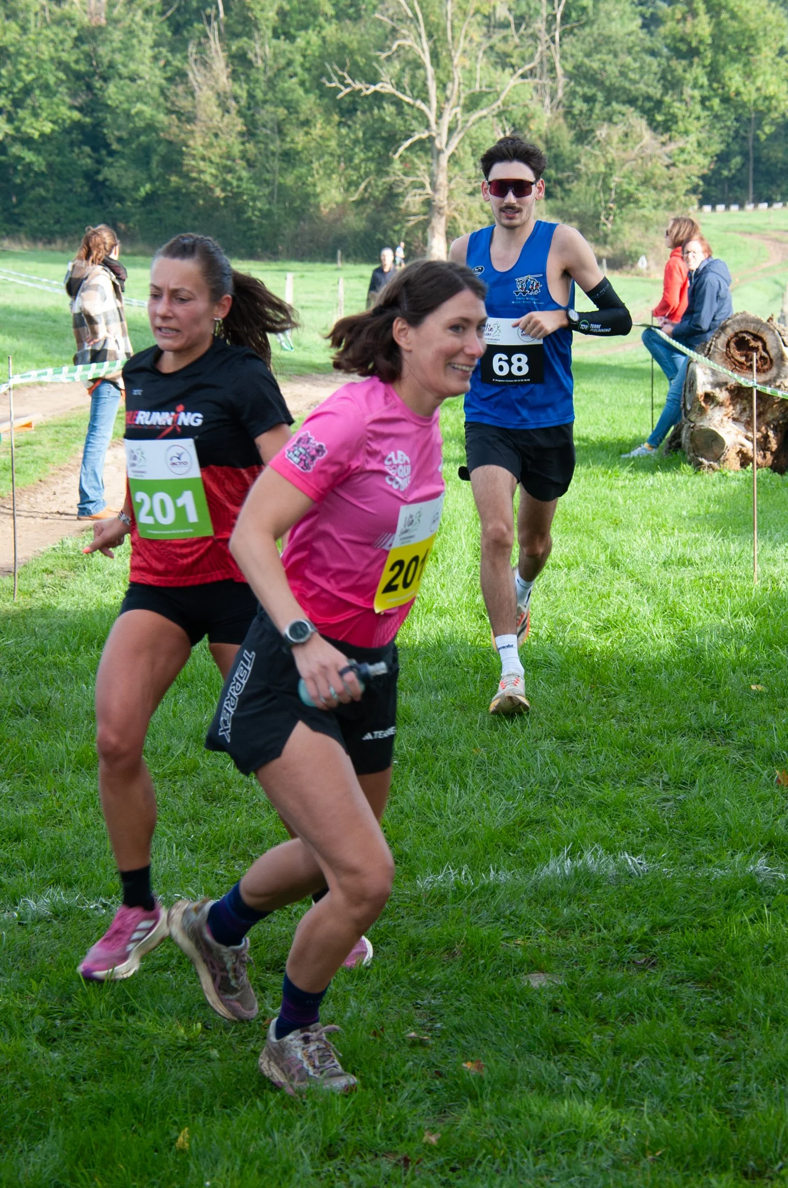 Trois coureurs féminines et un masculin participent à une course en pleine nature, avec des spectateurs en arrière-plan.