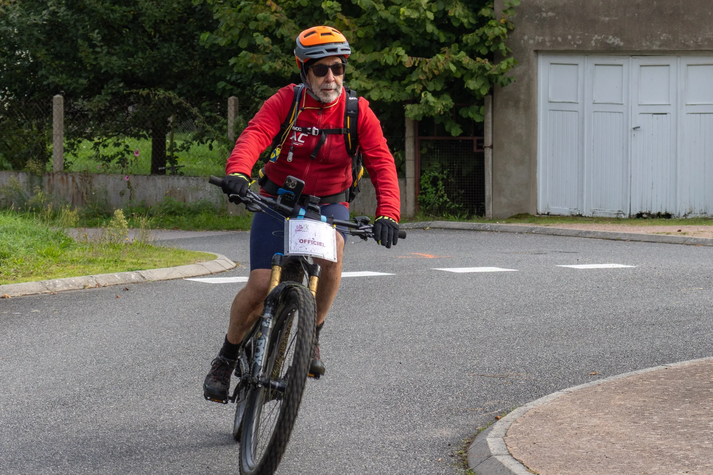 Un homme âgé portant un casque orange, des lunettes de soleil, une veste rouge, et des gants noirs, fait du vélo sur une route. Il porte un sac à dos et un bracelet avec une petite tablette attachée au guidon. Le fond montre un bâtiment avec des port