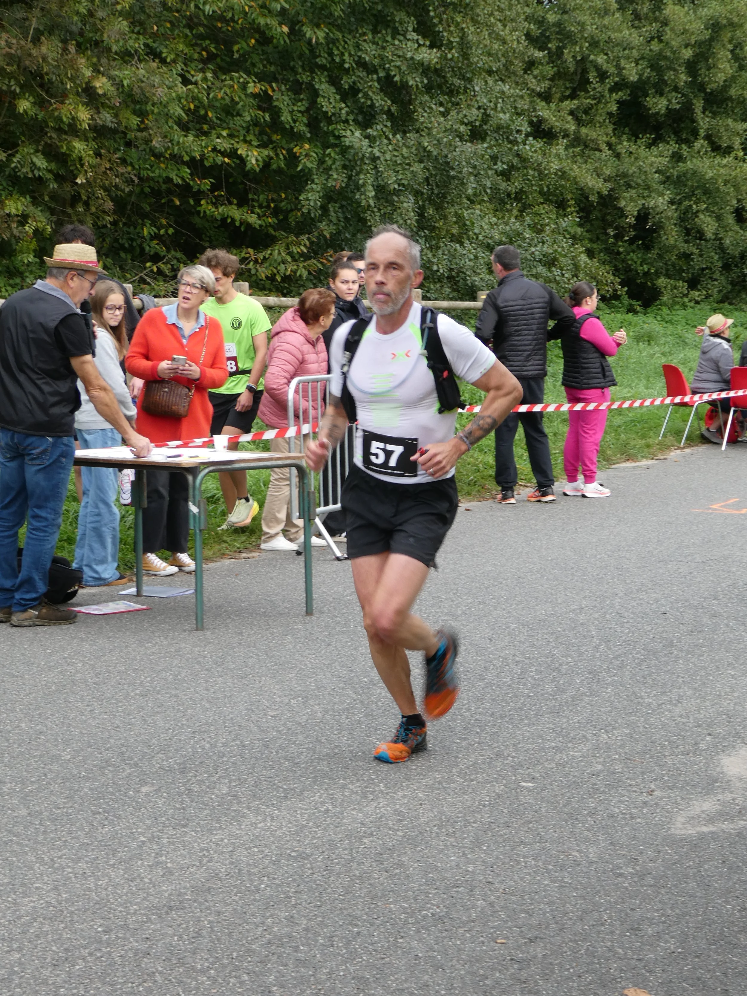 Un homme en course lors d'une compétition de course ou de marathon, portant un t-shirt blanc, un casque de course, un dossard numéro 57, et un sac à dos, avec des spectateurs et des bénévoles en arrière-plan.