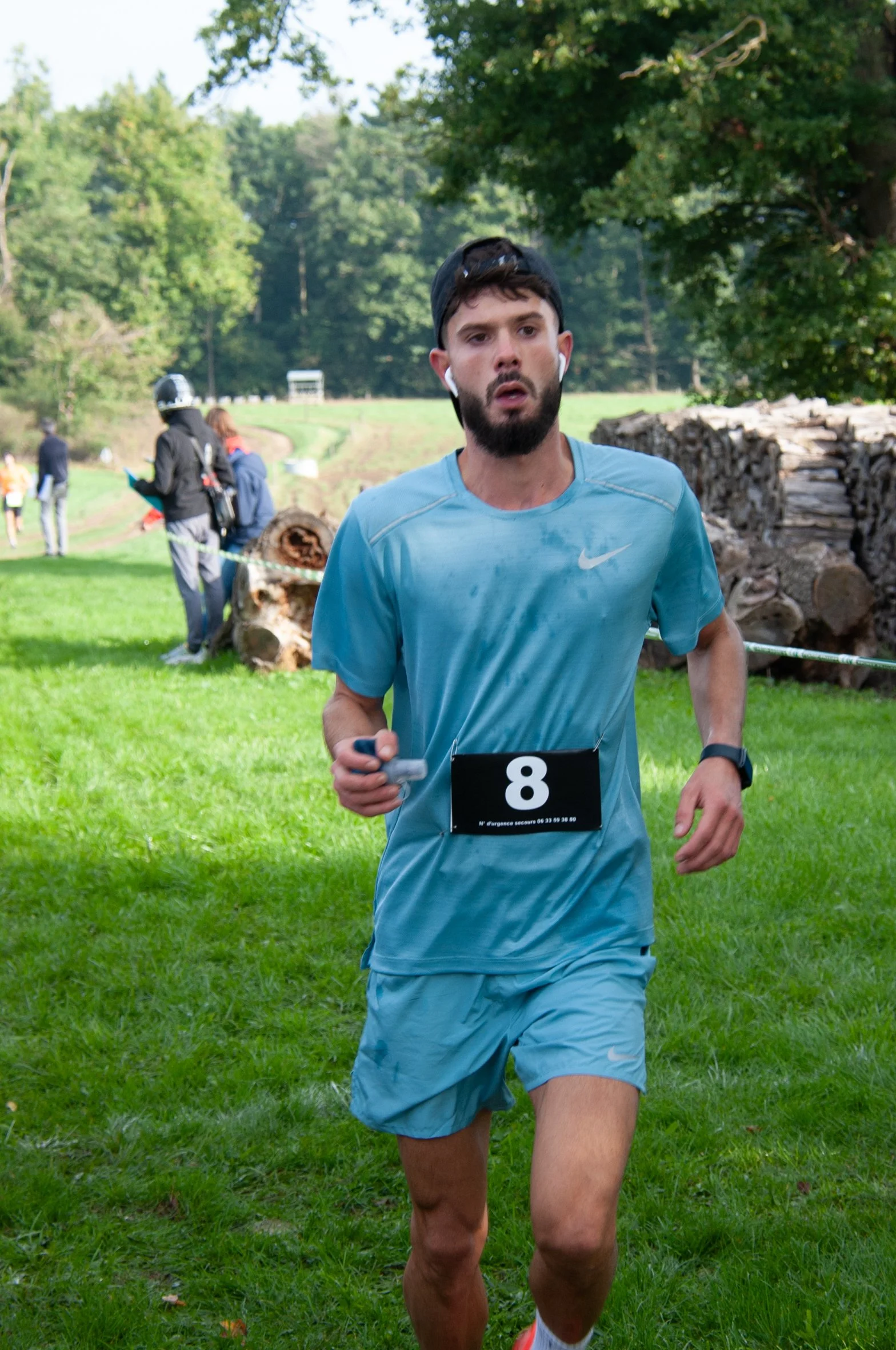 Un homme en tenue de course à pied, portant un numéro 8, court dans un parc vert entouré d'arbres, avec d'autres personnes en arrière-plan, certains regardant ou préparés pour une course.