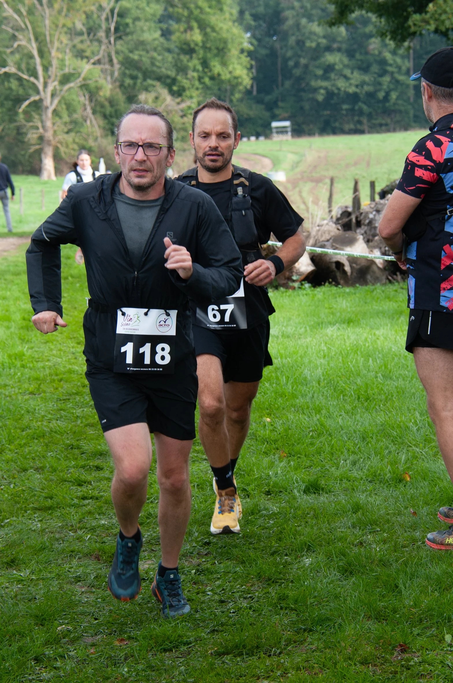Deux hommes en train de courir dans un champ lors d'une course, portant des dossards numérotés 118 et 67, avec un paysage verdoyant en arrière-plan.