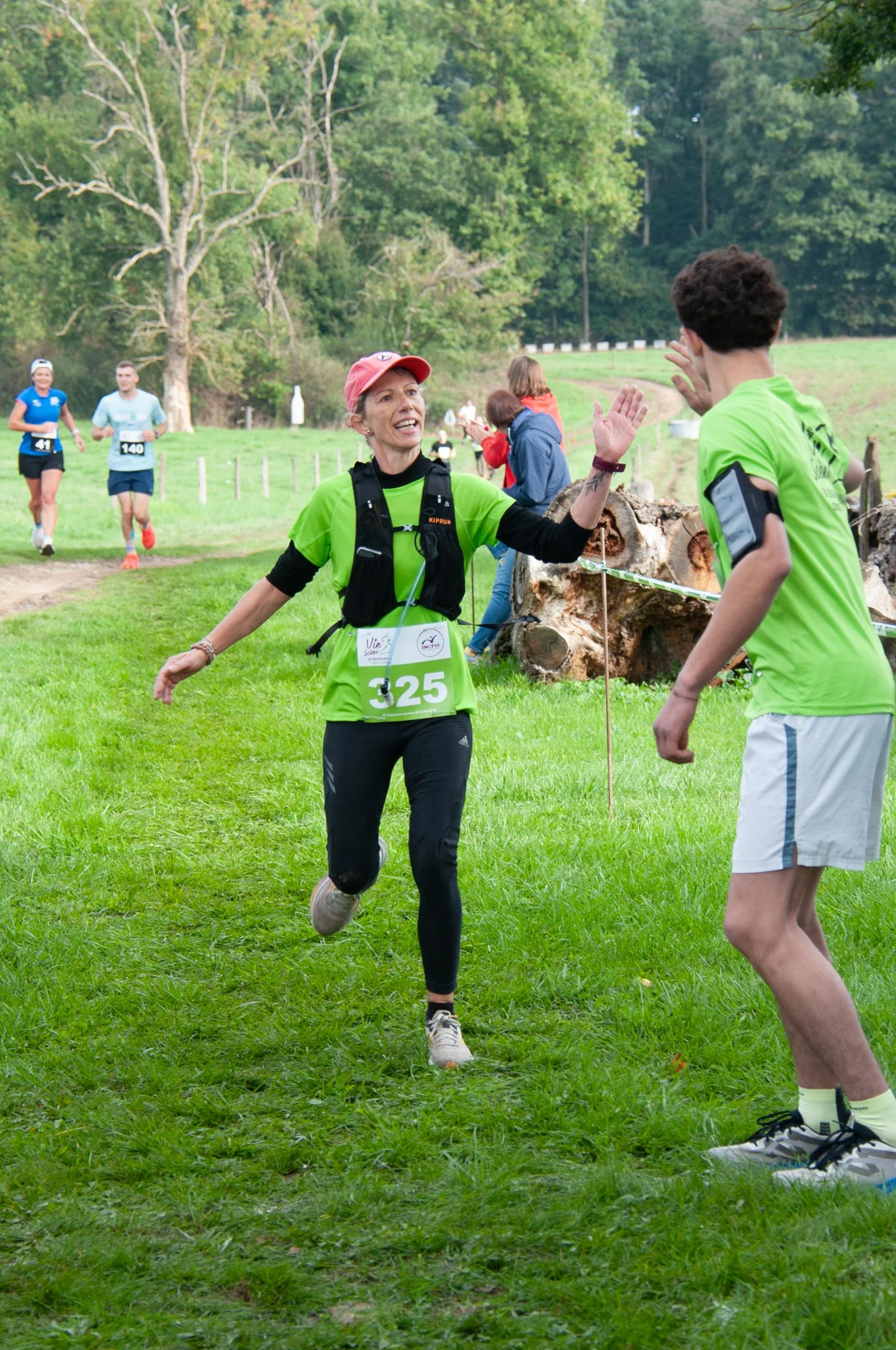 Une femme en course à pied dans un parc, souriante, échangeant un sourire avec un jeune homme. Elle porte un t-shirt vert, un rameur de course, un numéro de dossard 325, et des chaussures de sport. En arrière-plan, d'autres coureurs participent à une