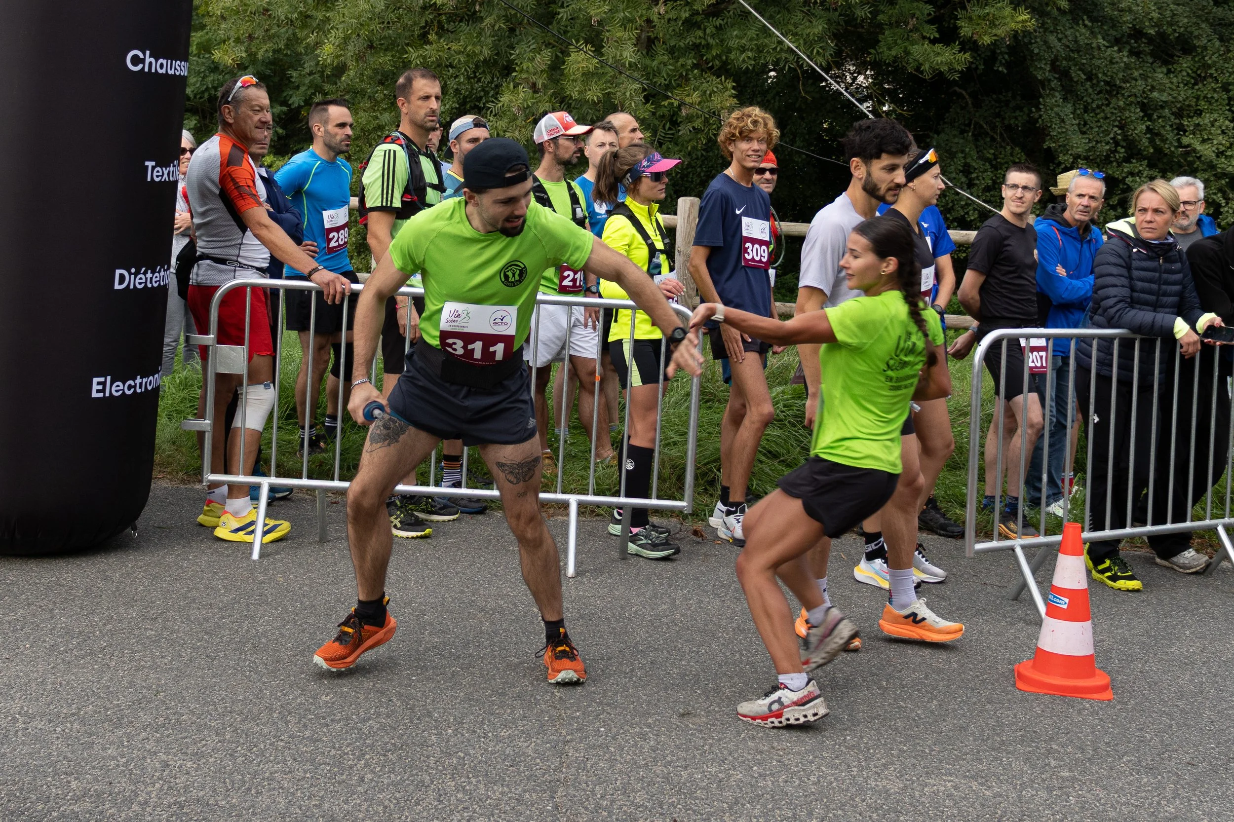 Un homme et une femme en tenue de course à l'extérieur, échangeant un high five près d'une barrière de course. Plus de coureurs et spectateurs sont en arrière-plan.