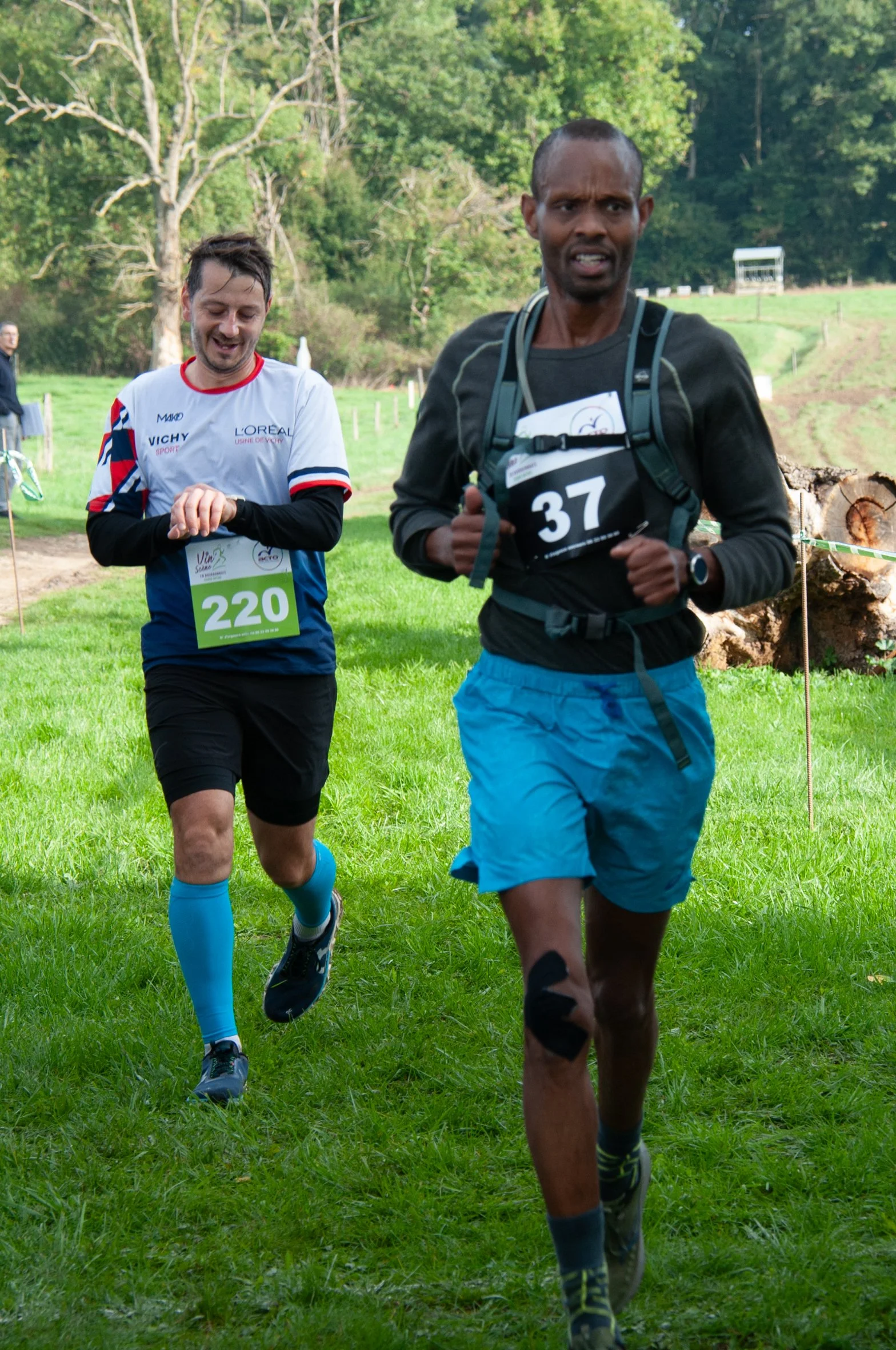 Deux coureurs participant à une course en plein air, portant des dossards numérotés 220 et 37, dans un environnement verdoyant avec des arbres en arrière-plan.