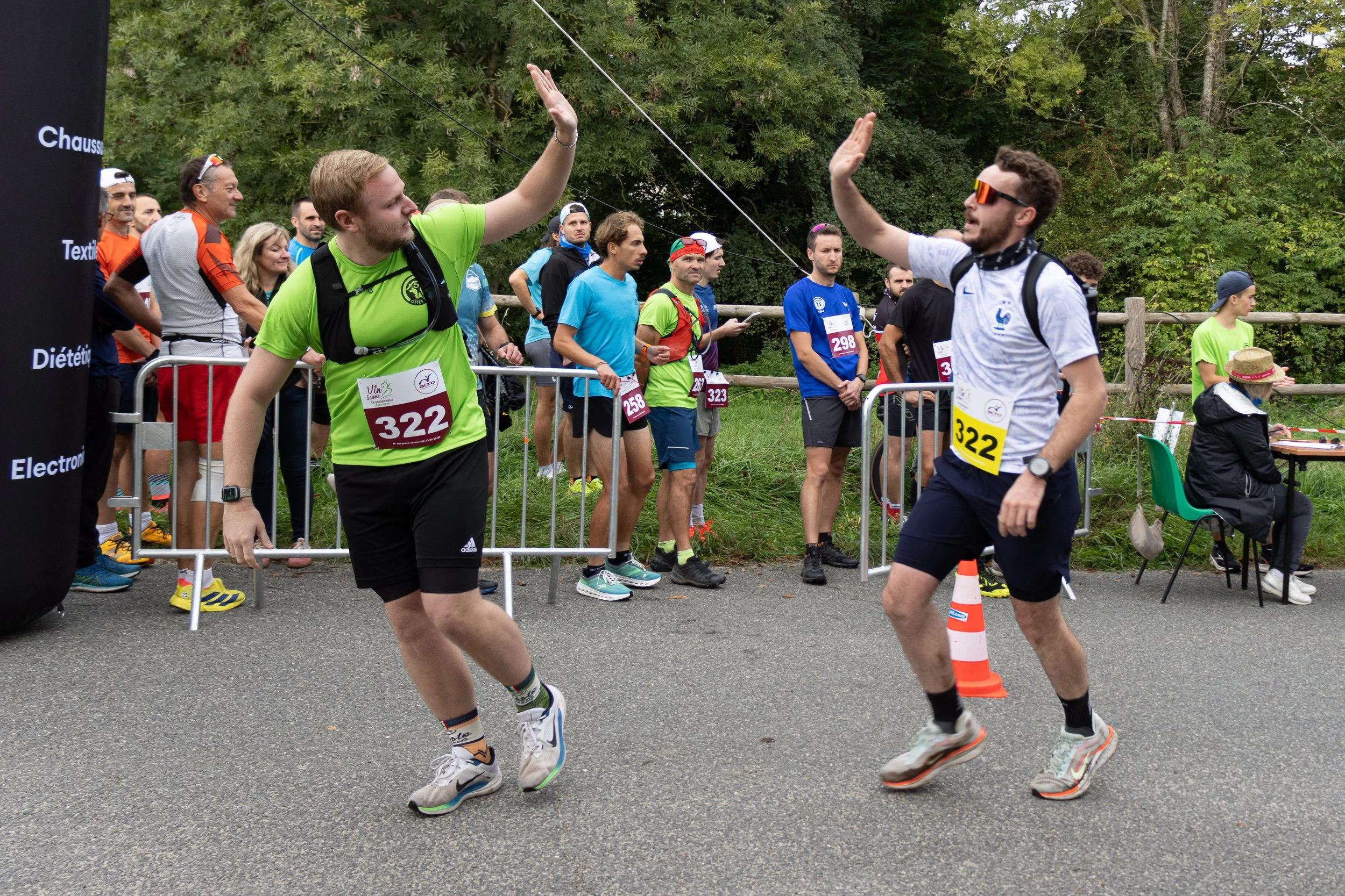 Deux coureurs se saluant avec des gestes de victoire lors d'une course, entourés de spectateurs et d'autres participants, en plein air, avec un fond de verdure.