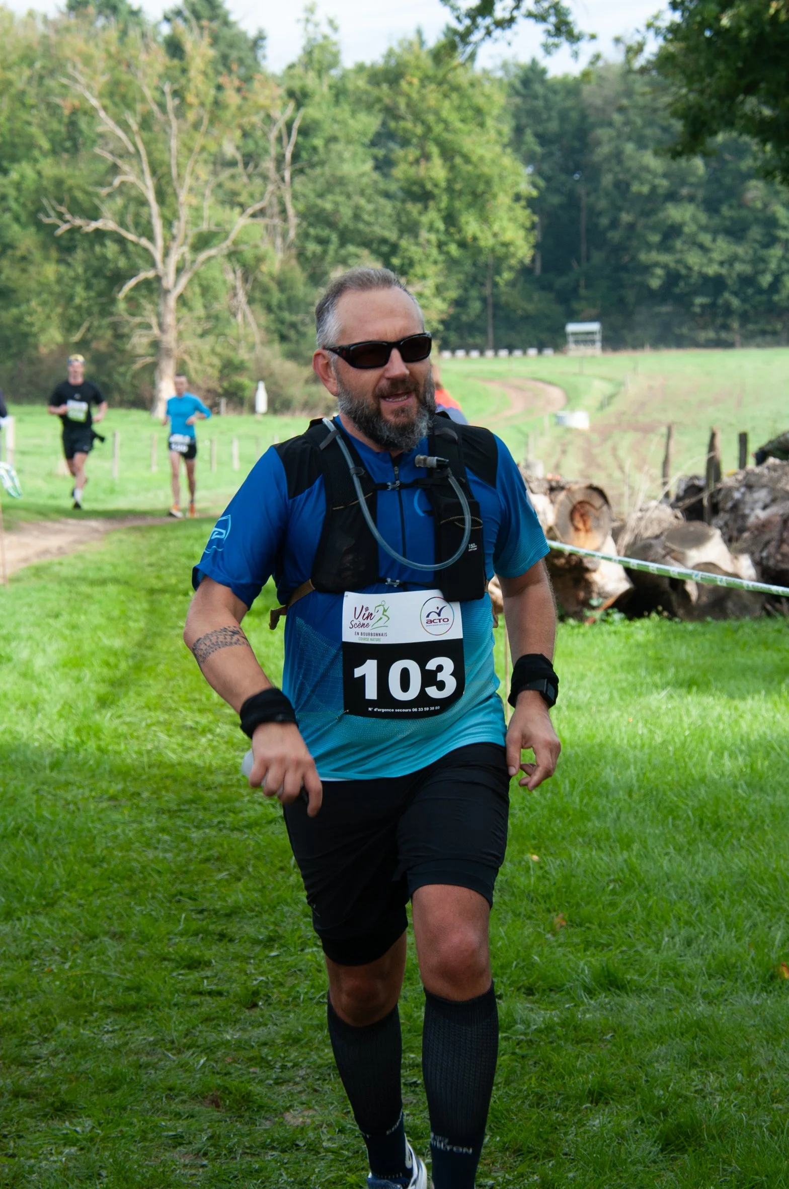 Homme courant dans une course en plein air avec un numéro 103, portant une tenue de course bleue, des lunettes de soleil, et un sac d'hydratation, dans un environnement naturel avec de l'herbe verte et des arbres.