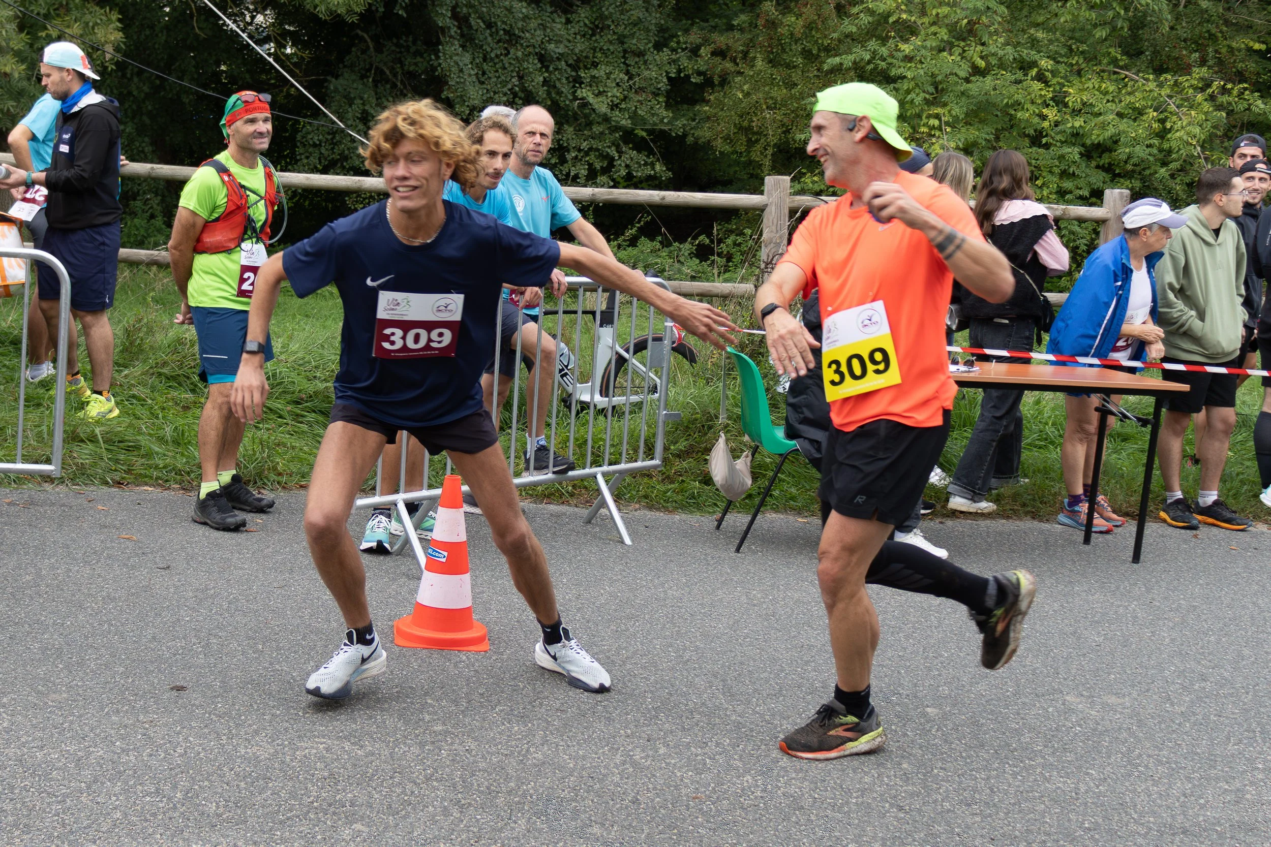 Deux coureurs commencent à se saluer à la fin d'une course de marathon, devant un groupe de spectateurs et de bénévoles. L'environnement est en plein air avec des arbres en arrière-plan.