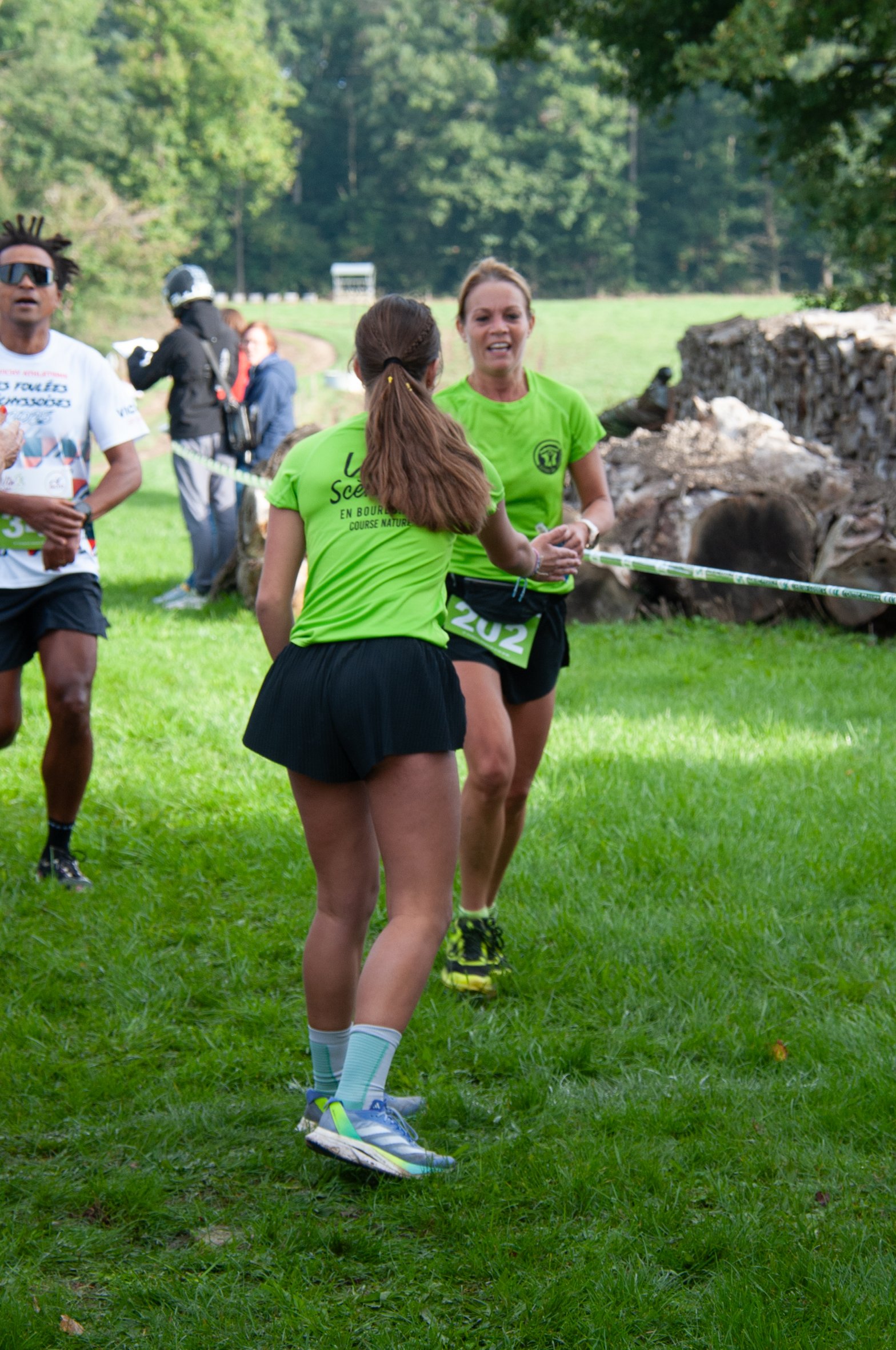 Deux femmes en tenue de course discutent durant une compétition en plein air, avec un terrain herbeux, des arbres et quelques spectateurs en arrière-plan.