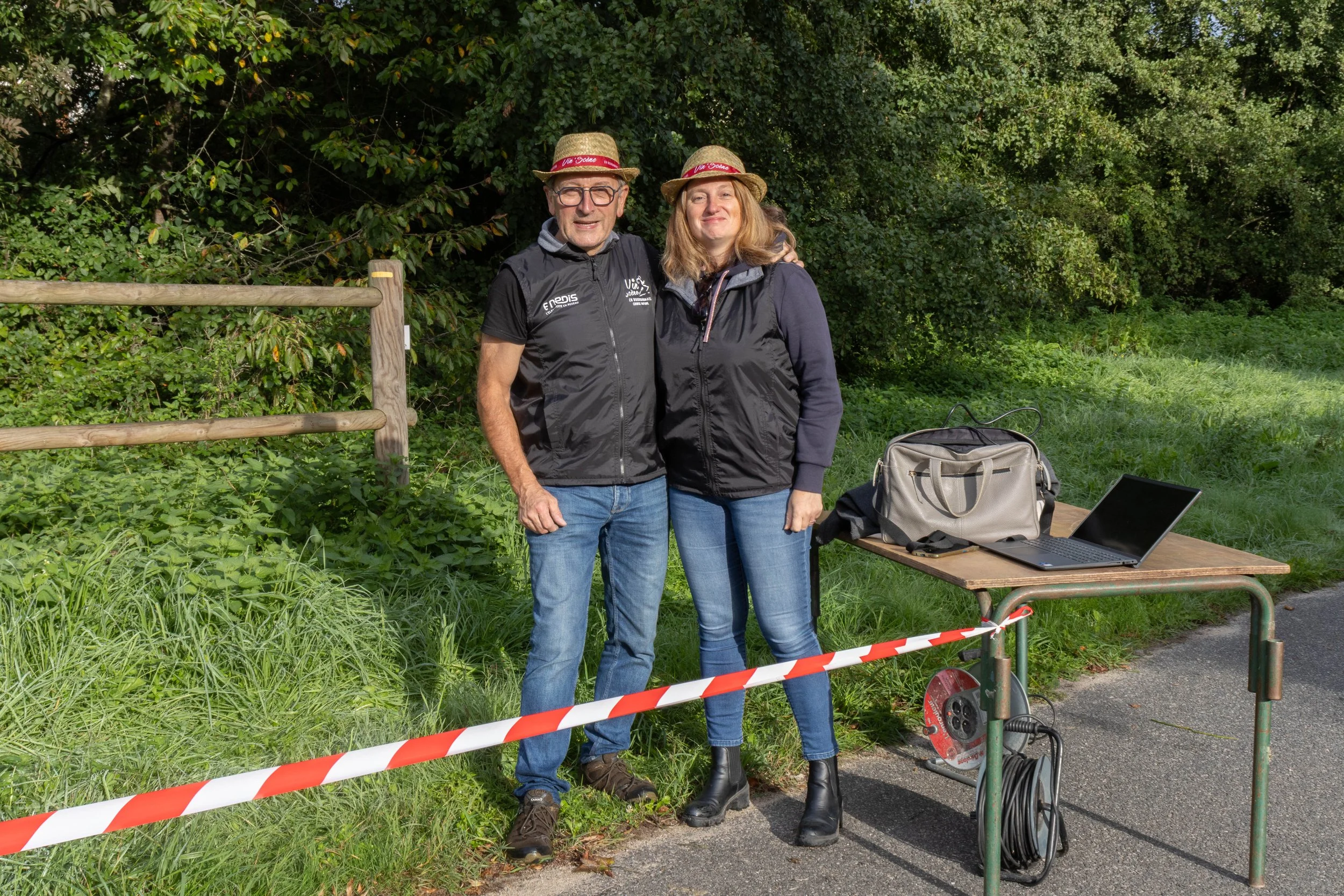 Deux personnes debout près d'une table en extérieur, un homme et une femme, souriants, portant des chapeaux en paille. La table a un ordinateur portable et un sac. La scène est en plein air avec des arbres en arrière-plan.