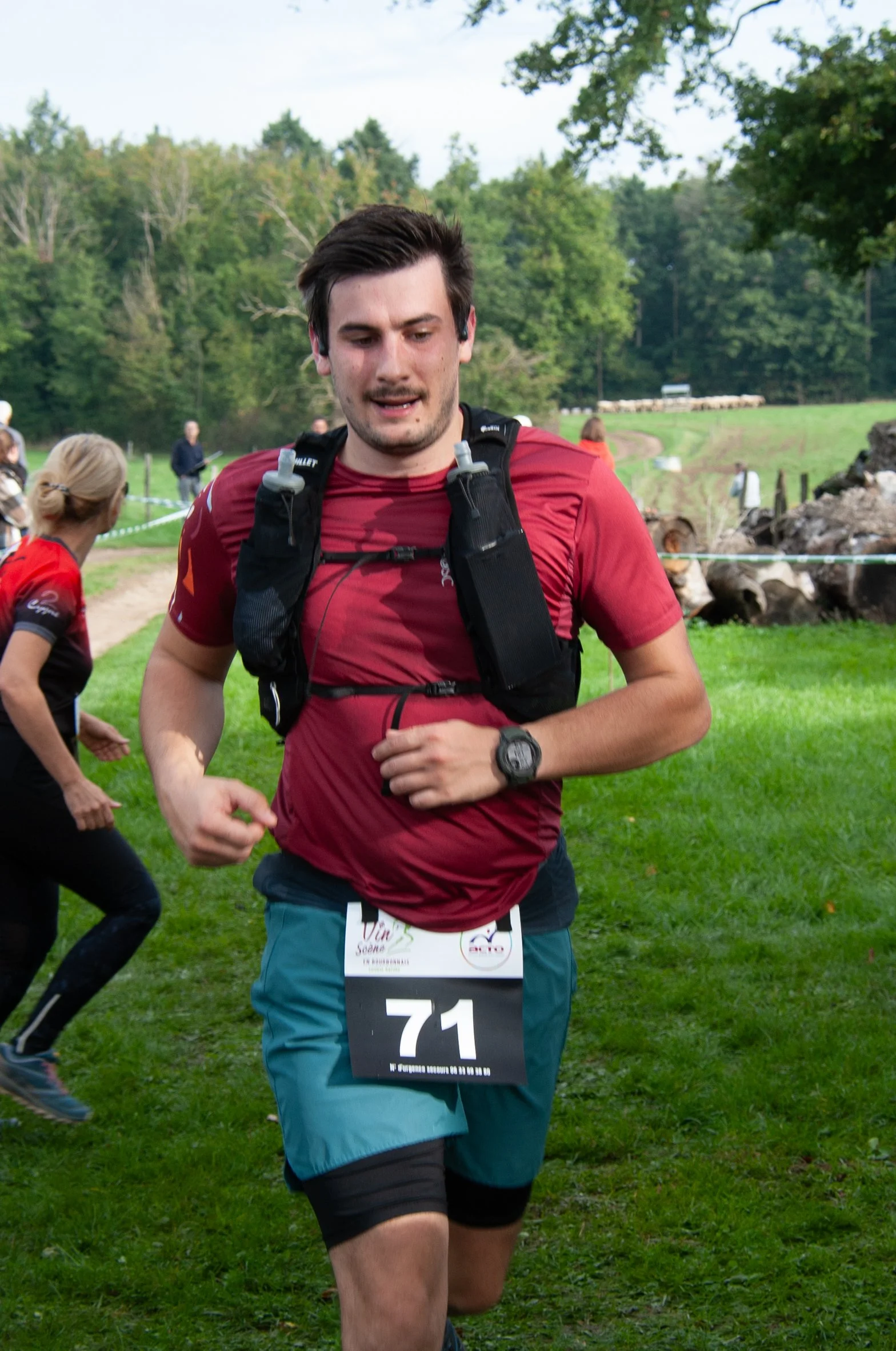 Un homme courait lors d'une course en plein air, portant un t-shirt rouge, un short bleu et un bracelet connecté, avec un numéro 71 attaché à sa taille, dans un environnement verdoyant avec d'autres participants en arrière-plan.