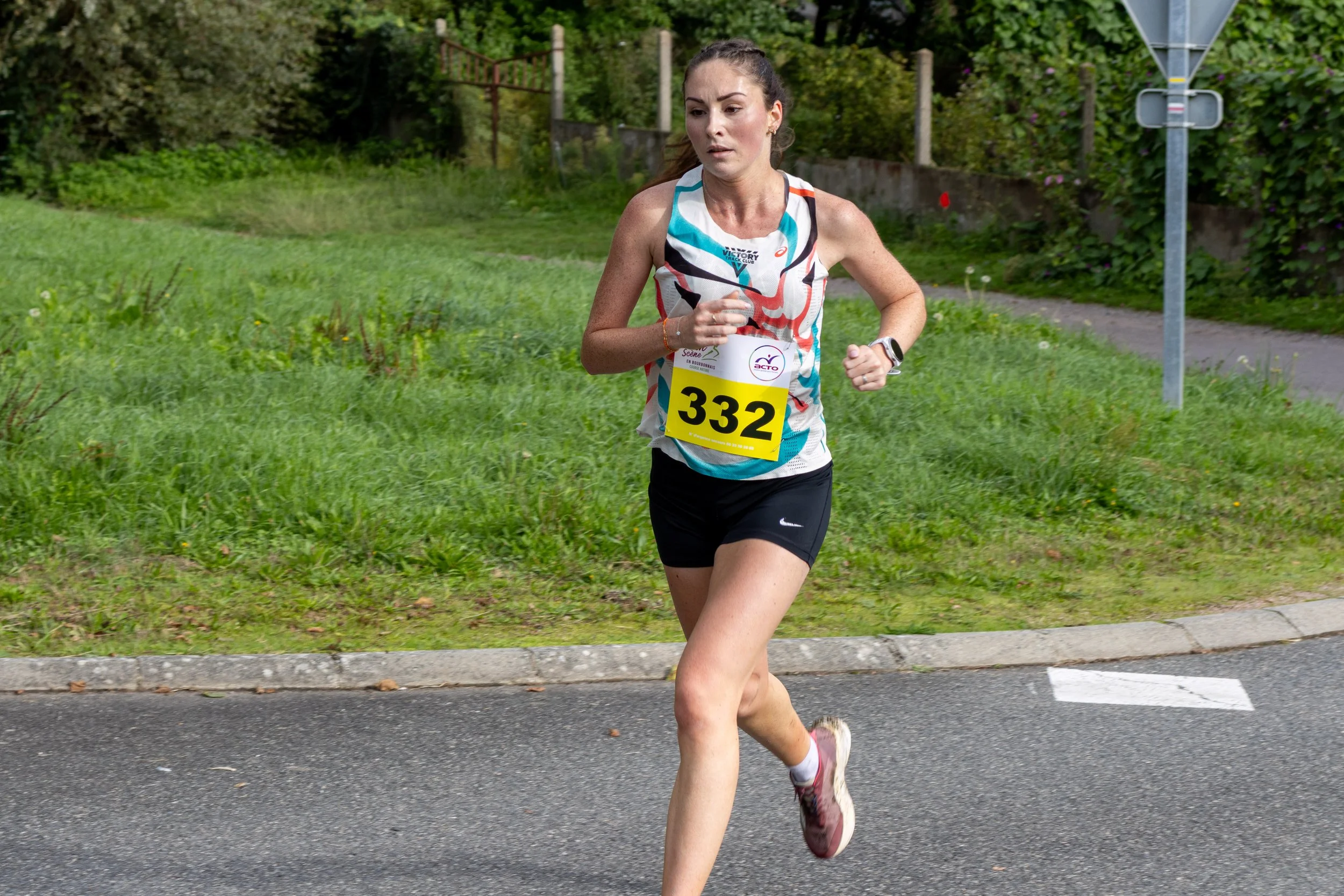 Femme athlète courant lors d'une course, portant un numéro 332, vêtue d'un débardeur coloré et d'un short noir, en pleine marche sur une route avec de la verdure en arrière-plan.