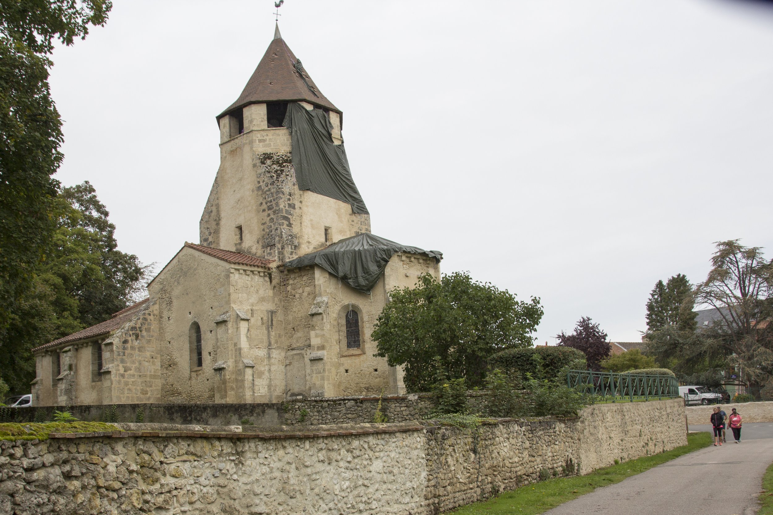 Église ancienne sous rénovation avec protection en plastique noire sur le clocher, entourée d'un mur en pierre, avec deux personnes marchant sur le chemin à droite.
